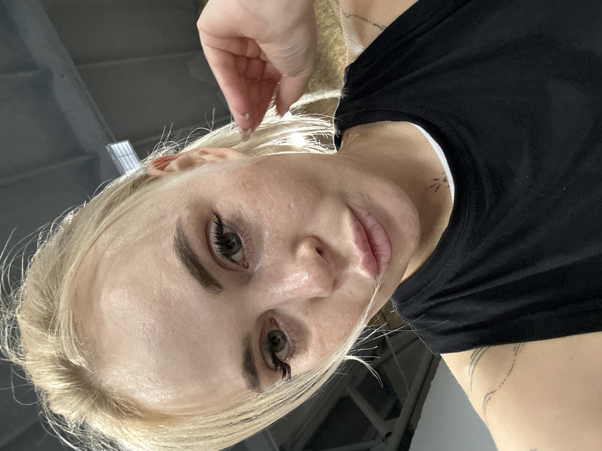 A close-up selfie shows a fair-skinned blonde woman with visible facial pores and dark mascara on her eyelashes. She is wearing a black t-shirt with a white trim at the neck. A hand is slightly visible near her forehead, and there are several tattoos on her neck and upper arm. The location appears to be indoors, possibly a utilitarian space, with a gray ceiling visible in the background. The image was potentially taken in Megion, Russia.