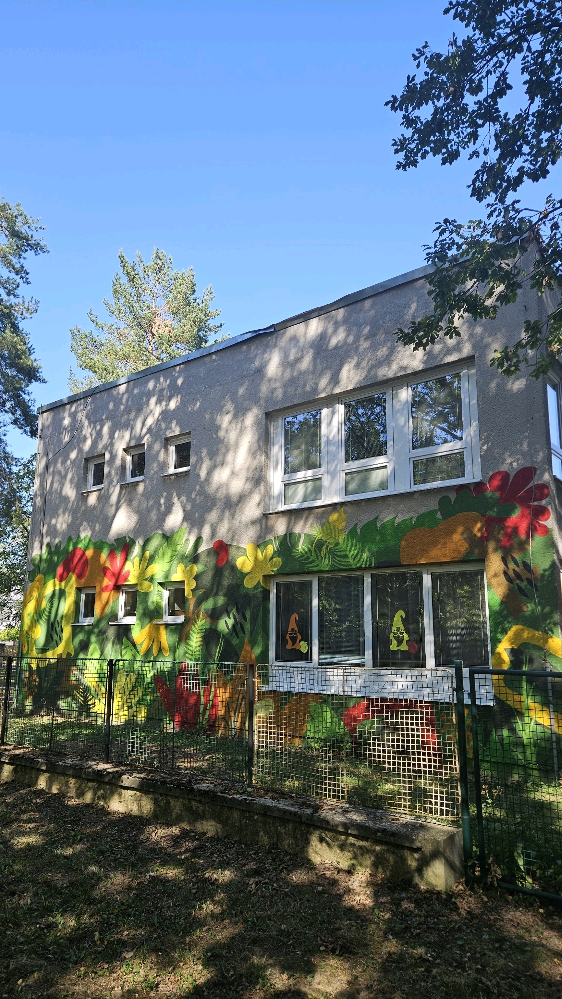 The image shows a one-story building with a colorful mural painted on its exterior wall, depicting jungle-like foliage and flowers. The mural wraps around the side of the building and features large, abstract shapes in red, yellow, green, and orange. Two windows on the lower level of the building each have a sticker of an orange gnome with a red hat. The building has a light-colored concrete facade and several rectangular windows on both the upper and lower levels. A metal fence with a wire mesh runs along the front of the building, separating it from a grassy area with fallen leaves. Trees and branches are visible in the foreground and upper part of the image, casting shadows on the building and ground. The sky is clear and blue. The building is located at the kindergarten on Družicová Street in Prešov, Slovakia.