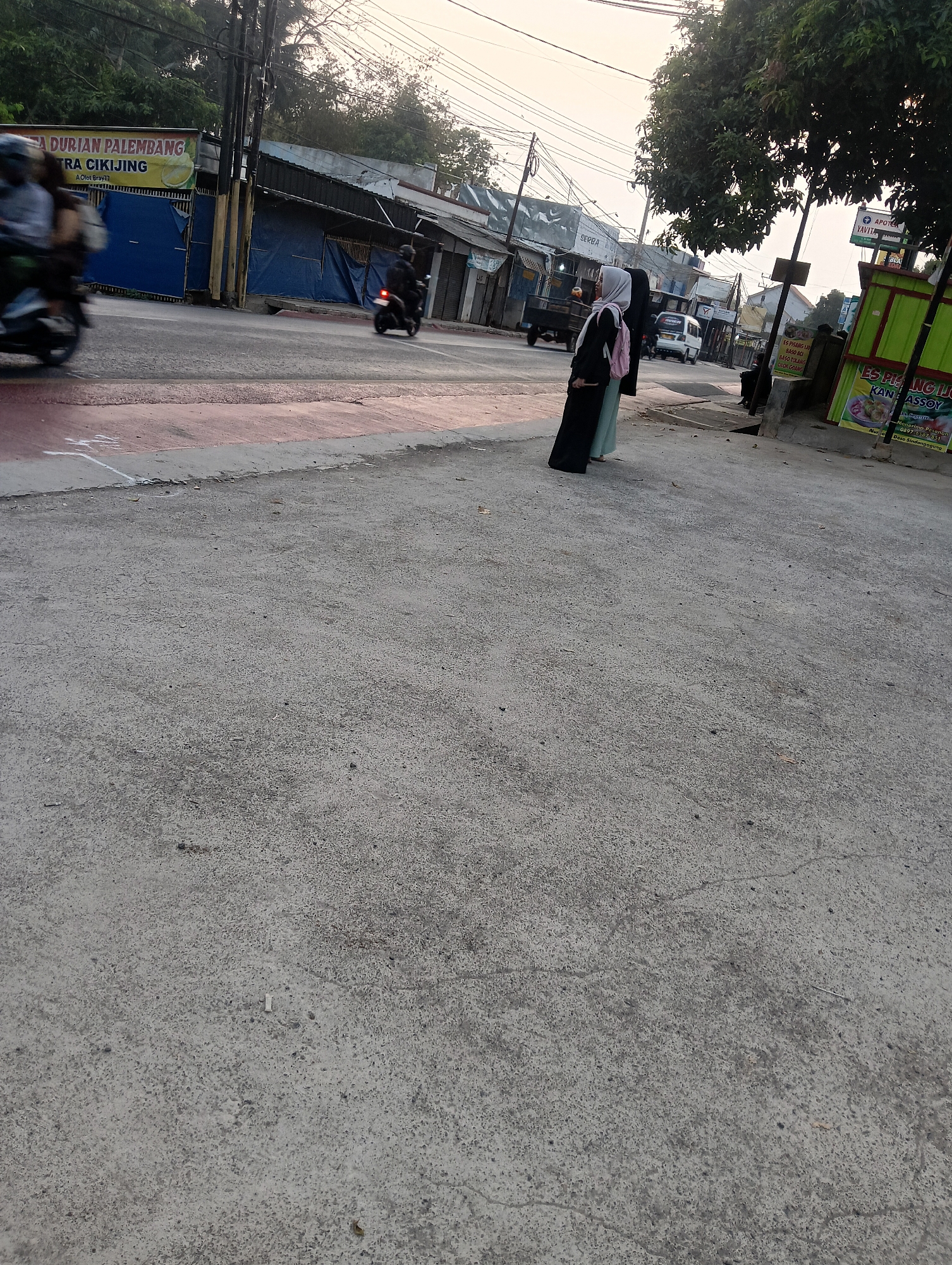 The image depicts a street scene in Manggari, Indonesia, with a focus on the pavement and background elements. In the foreground, rough, gray asphalt with visible cracks and small debris dominates the frame. To the left, a portion of a street with a reddish-pink lane marker is visible. In the middle ground, two women wearing hijabs stand on the sidewalk. One is wearing a black abaya and a light blue headscarf, while the other wears a light blue abaya and a grey headscarf. Behind them, a street with several vehicles, including a white van and a truck, is visible. A billboard advertising "DURIAN PALEMBANG" and "TRA CIKIJING" is also present. The background shows buildings with corrugated metal roofs and electrical wires, typical of urban Indonesian environments. The lighting suggests it is either early morning or late afternoon.