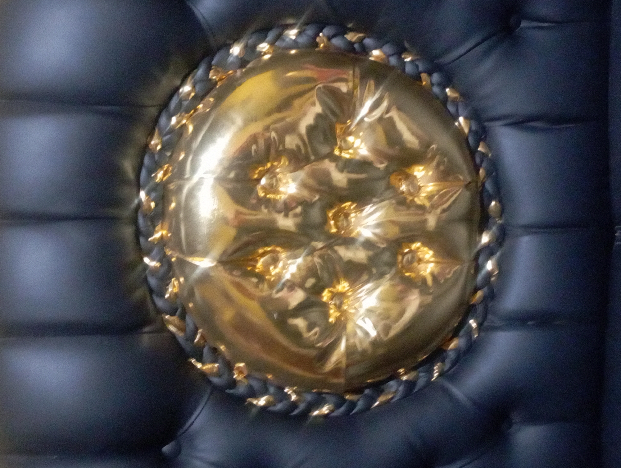 A close-up, eye-level shot captures the intricate details of a golden, tufted pillow set against a dark, button-tufted background. The pillow, circular in shape, features a textured braid or rope-like border, also in dark tones, interspersed with small, shiny gold accents. The surface of the golden pillow is highly reflective, showing distorted shapes and highlights that suggest it is made of a metallic or vinyl-like material. The background consists of what appears to be dark leather or faux leather upholstery, organized into regularly spaced square or rectangular tufts. The lighting is direct and dramatic, creating strong highlights and shadows on the pillow's surface, emphasizing its depth and texture. There are no people or discernable activities visible. The setting does not provide any specific clues to its location within Maos Kidul, Indonesia.