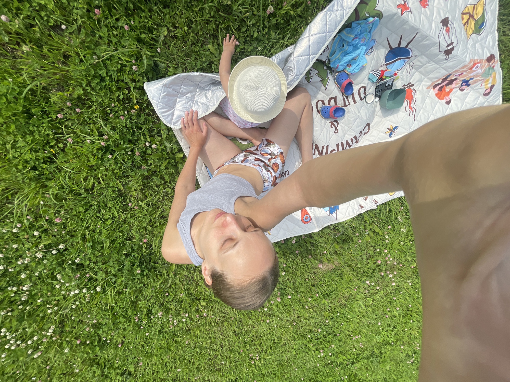 An overhead shot captures a woman and a baby sitting on a picnic blanket in a grassy field dotted with small pink and white flowers, presumably in or near the village of Podshivalovo, Russia. The woman, wearing a grey tank top and patterned shorts, is positioned on the left, with her eyes closed and head tilted to the side, appearing relaxed. Her left arm is extended, holding a camera to take the selfie. The baby, in front of the woman, is wearing a light-colored hat and a pink and purple outfit, with their head turned away from the camera. The picnic blanket displays cartoon graphics of camping scenes, including a family, a campfire, and a tent. A pair of blue children's clogs and a blue garment are also visible on the blanket. The background consists of lush green grass.