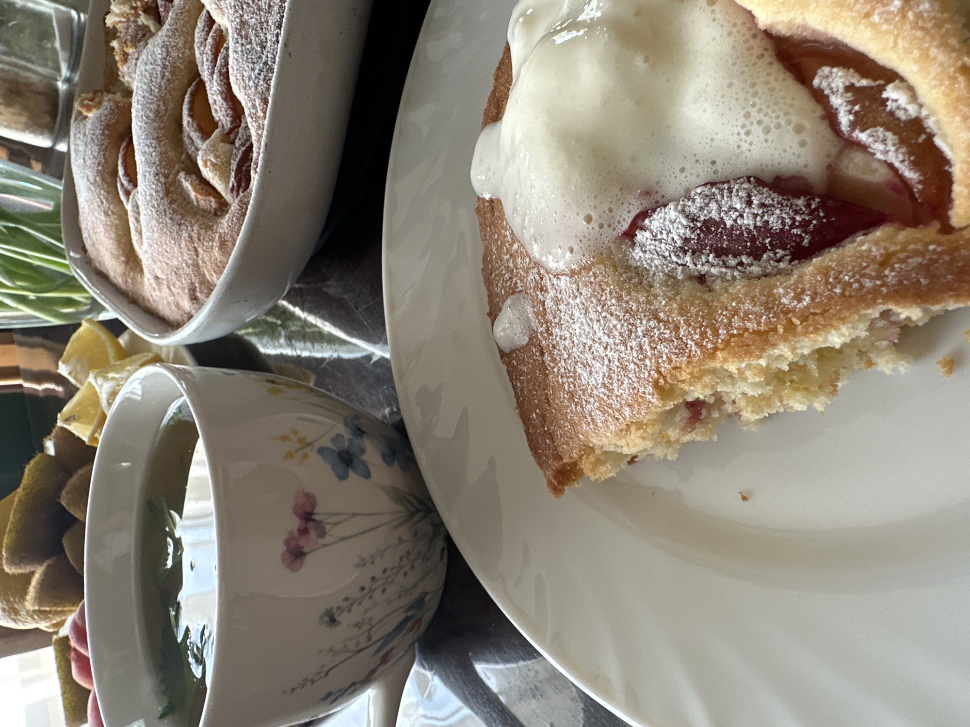The image presents a table setting with elements of a breakfast or brunch. A piece of baked cake, topped with what appears to be whipped cream and dusted with powdered sugar, is placed on a white plate. Beside it is a cup of tea in a floral-patterned teacup. Adjacent to the plate and cup is a baking dish filled with a similar cake, possibly a larger portion of the same cake on the plate. Green chives are visible, along with a lemon wedge. The overall setting suggests a home environment, likely indoors. There's no visible evidence that the scene is in Chaikovsky, Russia.