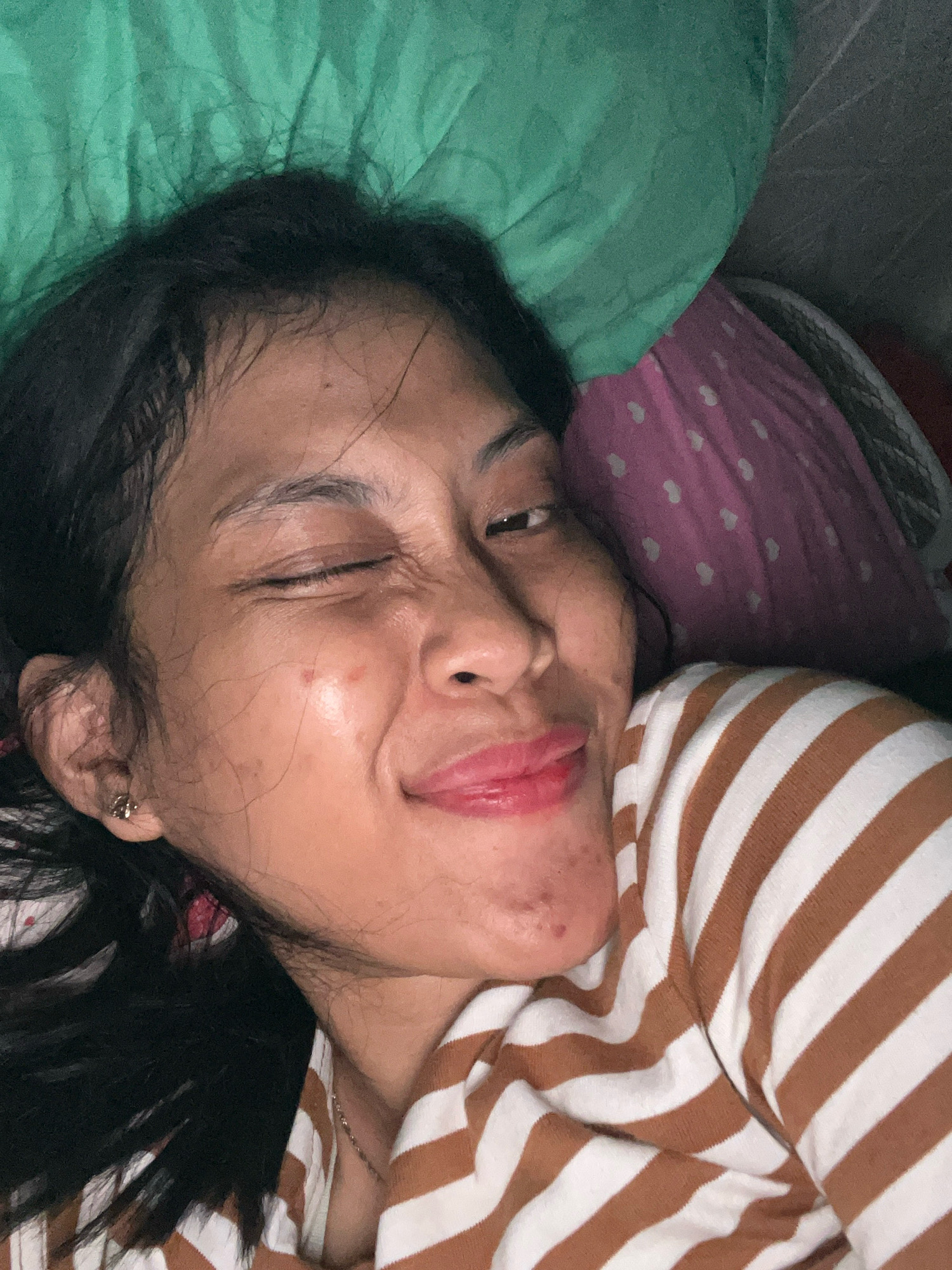 A woman lies on her side, looking at the camera with one eye closed in a wink. She has dark hair and is wearing a brown and white striped shirt. Her face shows some blemishes. She is on a bed with a green blanket and a pink pillow with white hearts. The scene appears to be indoors, likely in a bedroom, and there are no visible indicators of the specific location within Kecamatan Pati, Indonesia.