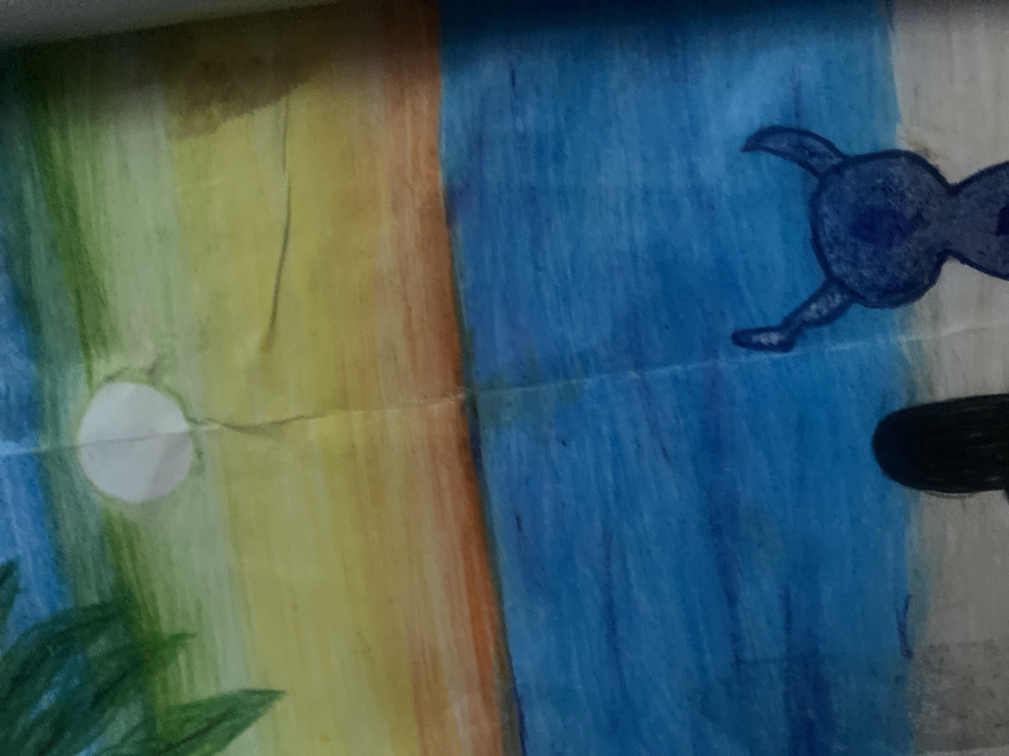 The image shows a drawing on paper, depicting two distinct scenes split vertically. On the left is a daytime scene with a yellow and orange sky, a white circle representing the sun or moon, and green foliage at the bottom. On the right is a blue scene with a dark blue, vaguely bird-shaped figure and a black oval shape. The paper has a crease running down the middle. The image does not include recognizable indicators of Ivaylovgrad, Bulgaria.