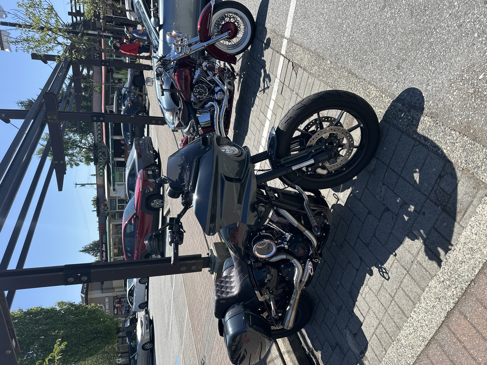 The image shows two motorcycles parked side-by-side on a brick-paved area next to a street. The motorcycle in the foreground is black and appears to be a modern Harley-Davidson bagger. The motorcycle behind it is red with whitewall tires and has more classic styling. Parked vehicles are visible on the street behind the motorcycles. Pedestrians are partially visible, suggesting a pedestrian zone or a busy street. Commercial buildings are in the background. The Dollar Store sign is visible, and this puts the location in Coquitlam, Canada. The sky is clear and blue.