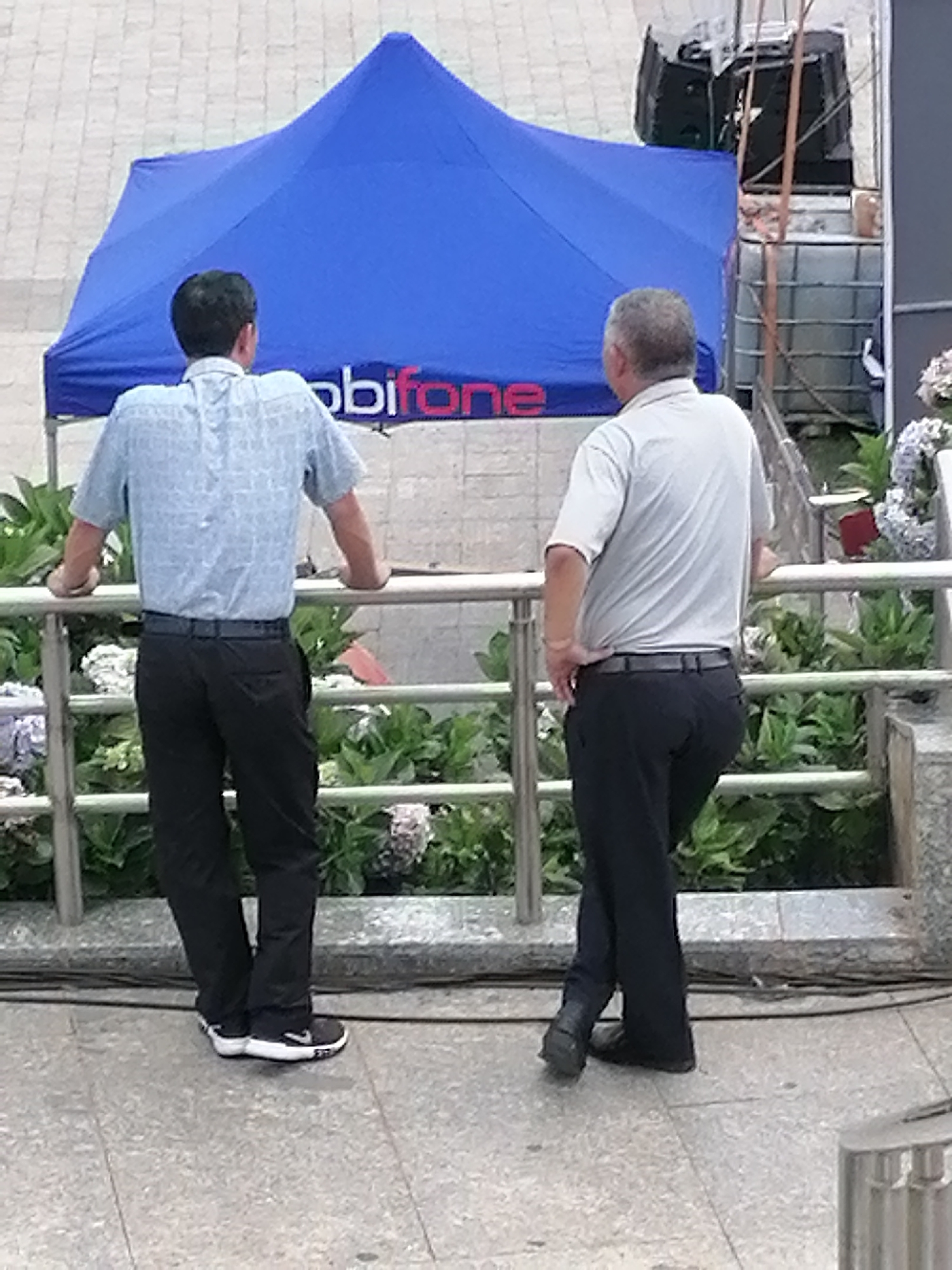 The image depicts two men standing near a blue event tent with the word "mobifone" visible on its side. The tent is situated on a paved outdoor area. The man on the left, seen from behind, wears a light blue patterned short-sleeved shirt and dark trousers, leaning with his hands on a metal railing. The man on the right, also seen from behind, wears a light-colored polo shirt and dark trousers, with his hands behind his back. Behind the men, there is lush green foliage and some white and purple flowers. In the background, there are industrial-looking containers and possibly equipment. The scene is likely a public space in Da Lat, Vietnam, possibly an outdoor event or market area, given the presence of the branded tent and the general urban setting.