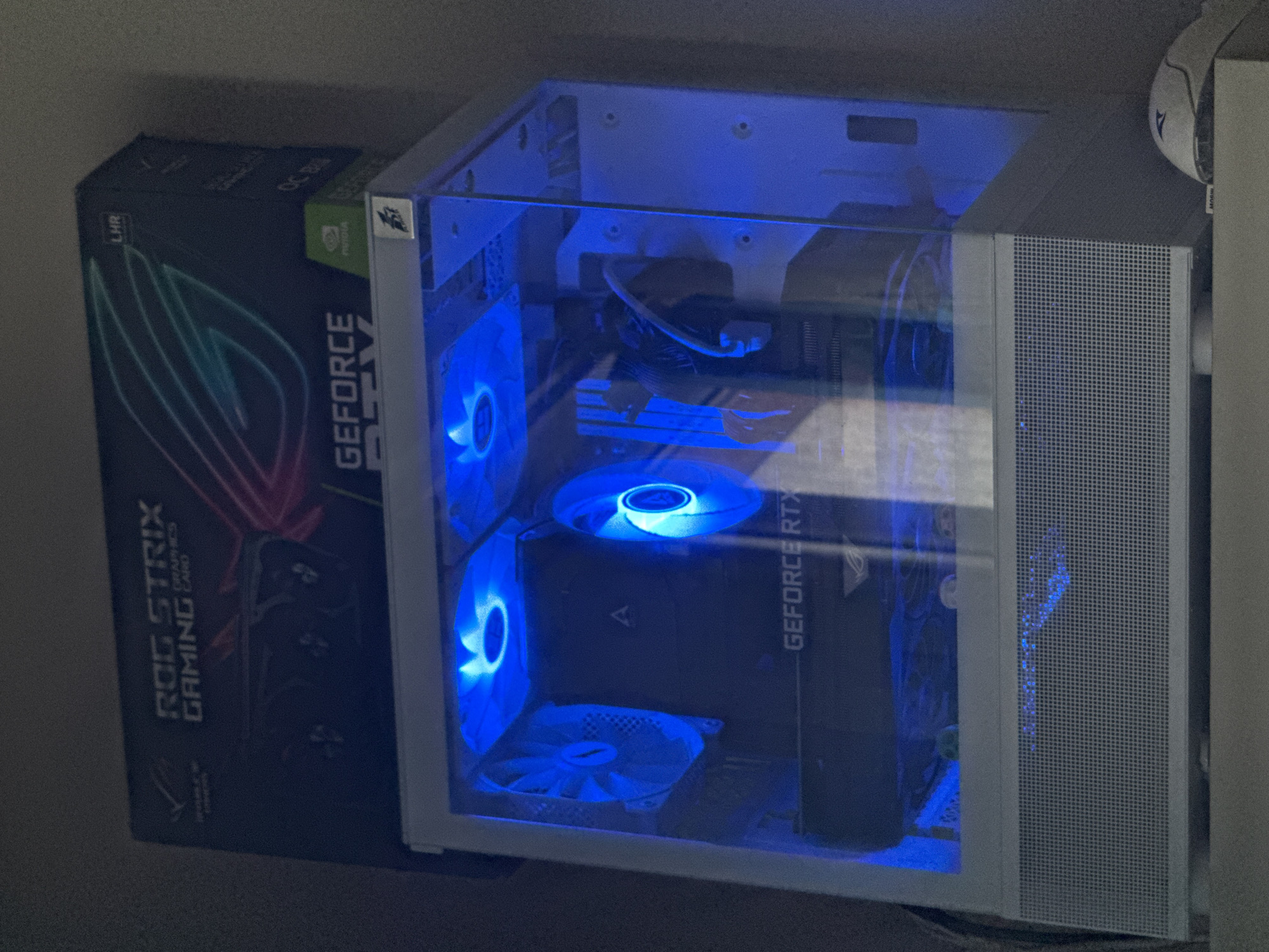 The image shows a computer case with a glass side panel revealing internal components illuminated by blue LED fans. A graphics card, labeled "GEFORCE RTX," is visible inside. The box for a ROG STRIX gaming graphics card is positioned next to the computer case. The environment is dimly lit. The specific location in Ruse, Bulgaria is not discernible from the image.