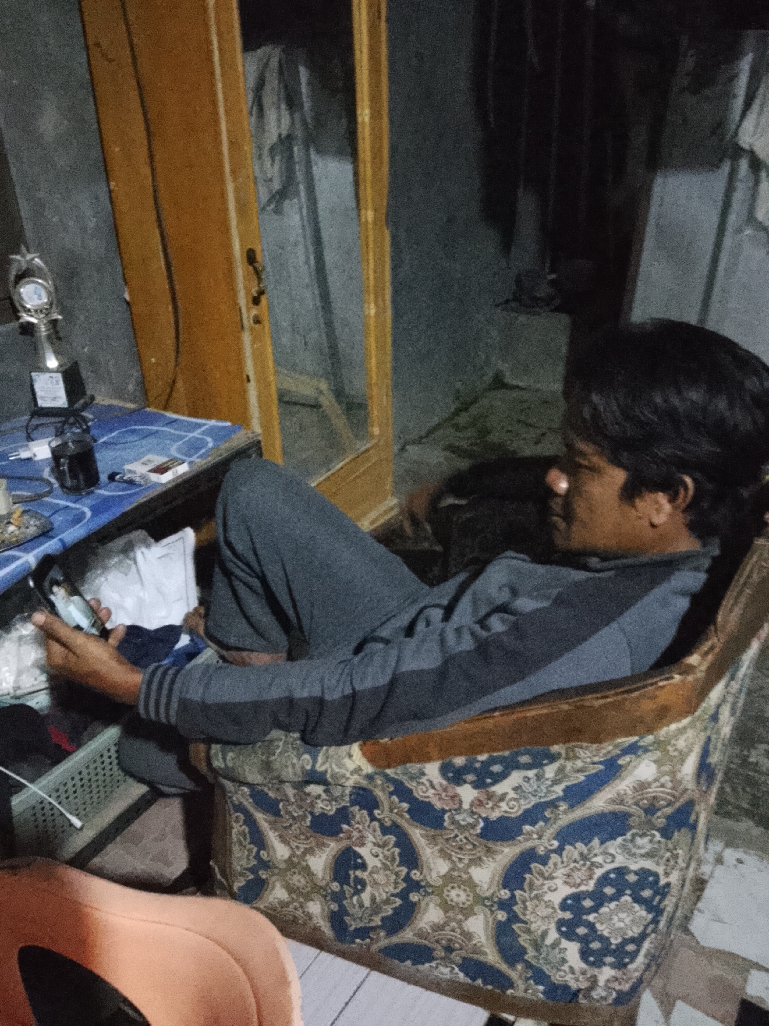 A man is sitting in a patterned armchair in a dimly lit room, holding a smartphone and looking at it. The room appears to be a residential dwelling. In the background, there is a wooden door frame and some shadows cast on the wall. On a blue table in the foreground, there are several objects including a trophy, a glass of dark liquid, a pack of cigarettes, and a white USB charging adapter. A white plastic basket is also visible. The man is wearing a grey sweatshirt with black stripes on the sleeves and grey pants. The armchair has a dark wood frame and is upholstered with a fabric featuring a blue and white floral pattern. The floor appears to be tiled. The scene is likely from Padang Dalom, Indonesia, as indicated by the image's metadata.