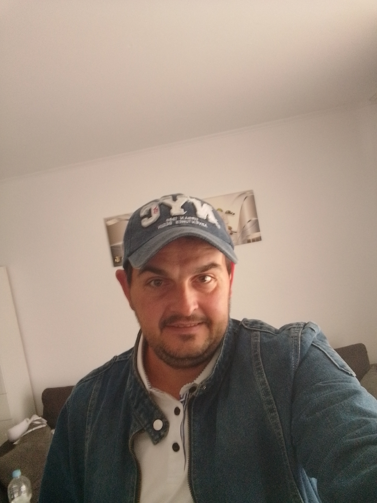 The image is a selfie of a man wearing a denim jacket and a blue baseball cap with white lettering that reads "NYC". He has dark hair, a mustache, and a light beard. The man is looking directly at the camera and smiling slightly. He is indoors, in what appears to be a living room. In the background, there is a white wall with a framed picture, a brown sofa, and a partially visible water bottle. The overall lighting is soft and diffused. The image does not provide any specific information about the location in the city of Wedel, Germany.