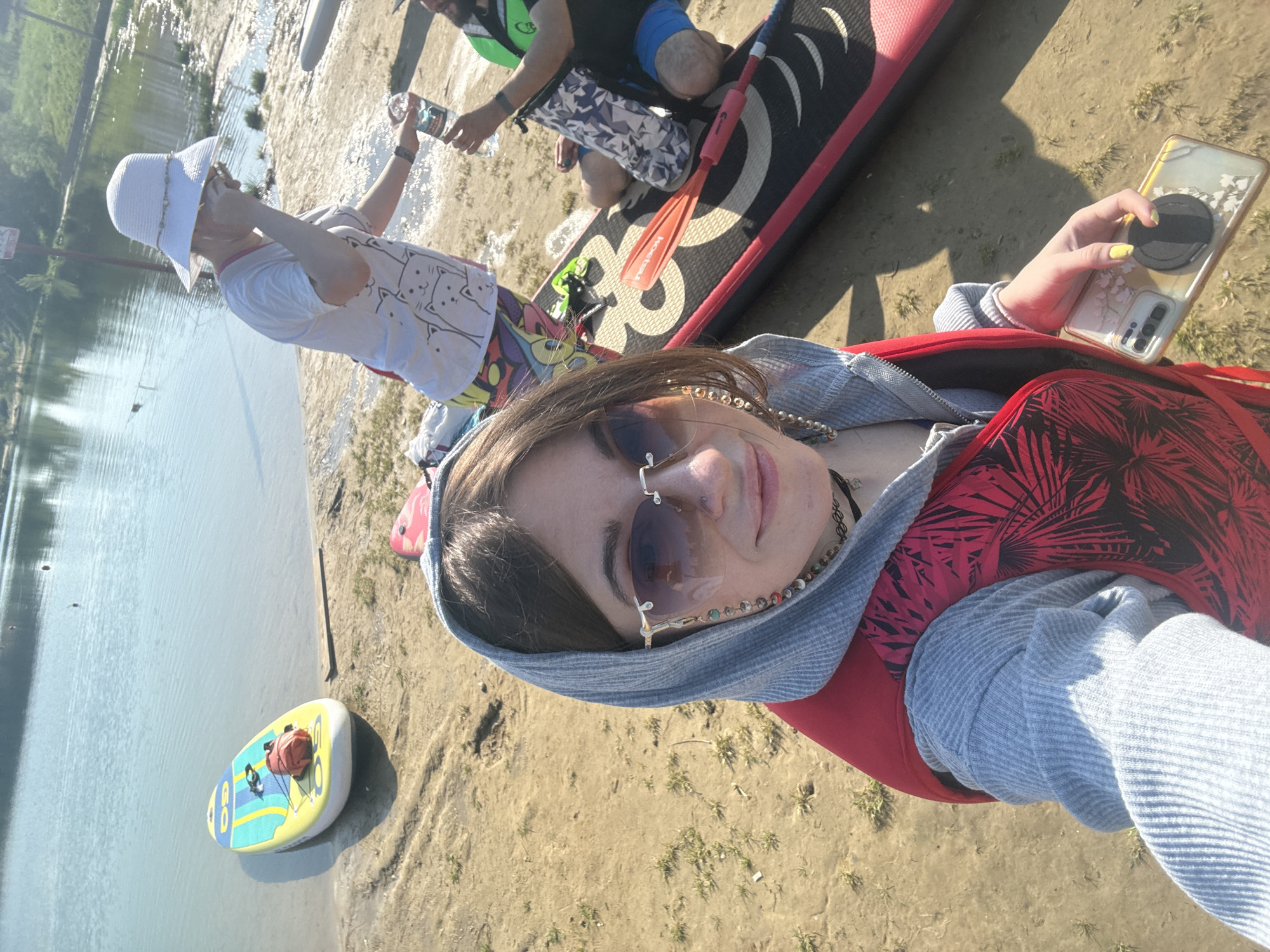 A woman wearing sunglasses, a red and black patterned vest, and a gray hoodie is taking a selfie on the sandy bank of a body of water. The bank is near the shore, where two other people stand beside a red and black paddleboard. One person is wearing a white hat and a light-colored shirt with cartoon cats on it. The other person is wearing a life vest and patterned shorts. A small inflatable raft is partially submerged in the water. The location is likely in Dzerzhinsky, Russia.