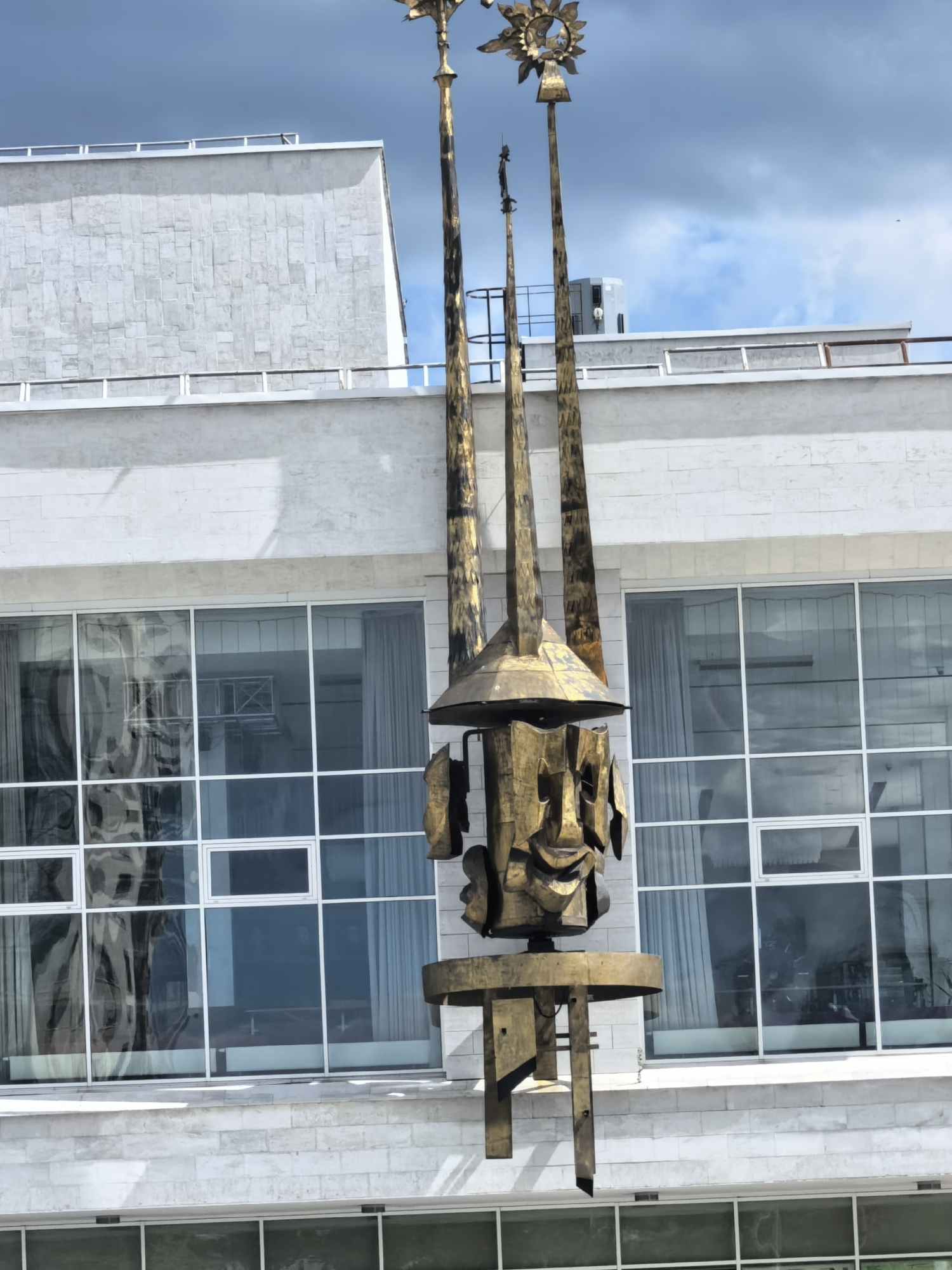The image shows a metallic sculpture positioned in front of a building. The sculpture consists of a multi-faceted, possibly humanoid figure beneath a conical roof, supported by vertical shafts. At the top of the shafts are decorative elements, including a sun-like figure. The building behind the sculpture has a modern design with large windows, reflecting interior elements and the outdoor environment. The building's facade is constructed of light-colored stone or brick, and there's a second level above with a balcony. The sky is visible above the building, showing a mix of dark, stormy clouds and patches of blue sky. The location is in the city of Unknown, Unknown.