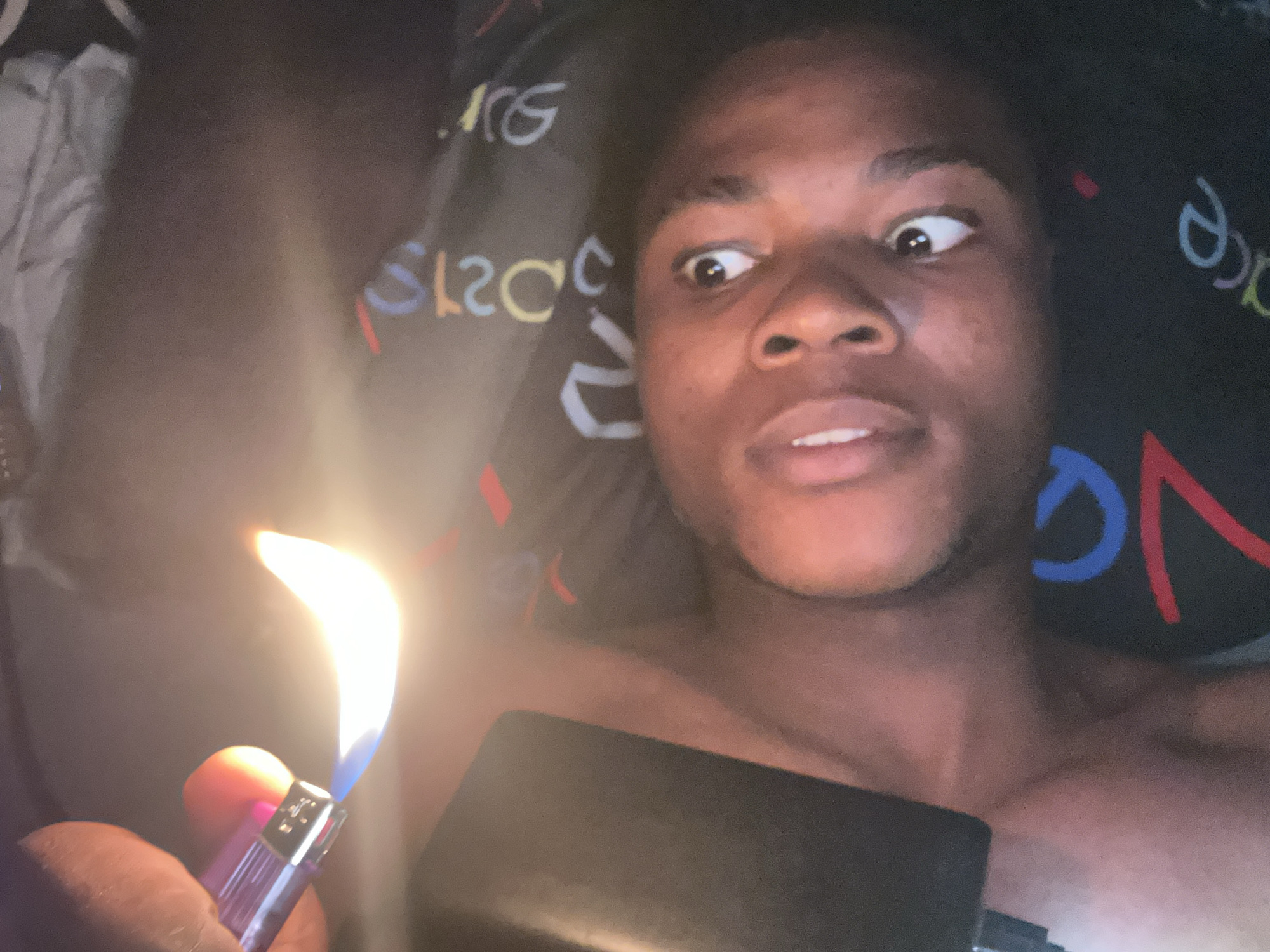 The image is a close-up, tilted shot of a person's face and hand holding a lighter with a visible flame. The person appears to be lying down, with part of their face illuminated by the lighter's glow. They have dark skin and their eyes are open, looking upwards. The background is a dark fabric with colorful lettering, possibly a t-shirt or blanket. The person's hand, which is holding the lighter, is also visible on the left side of the frame. The setting is indoor, and there is no clear indication of the specific location within Ihiagwa, Nigeria.