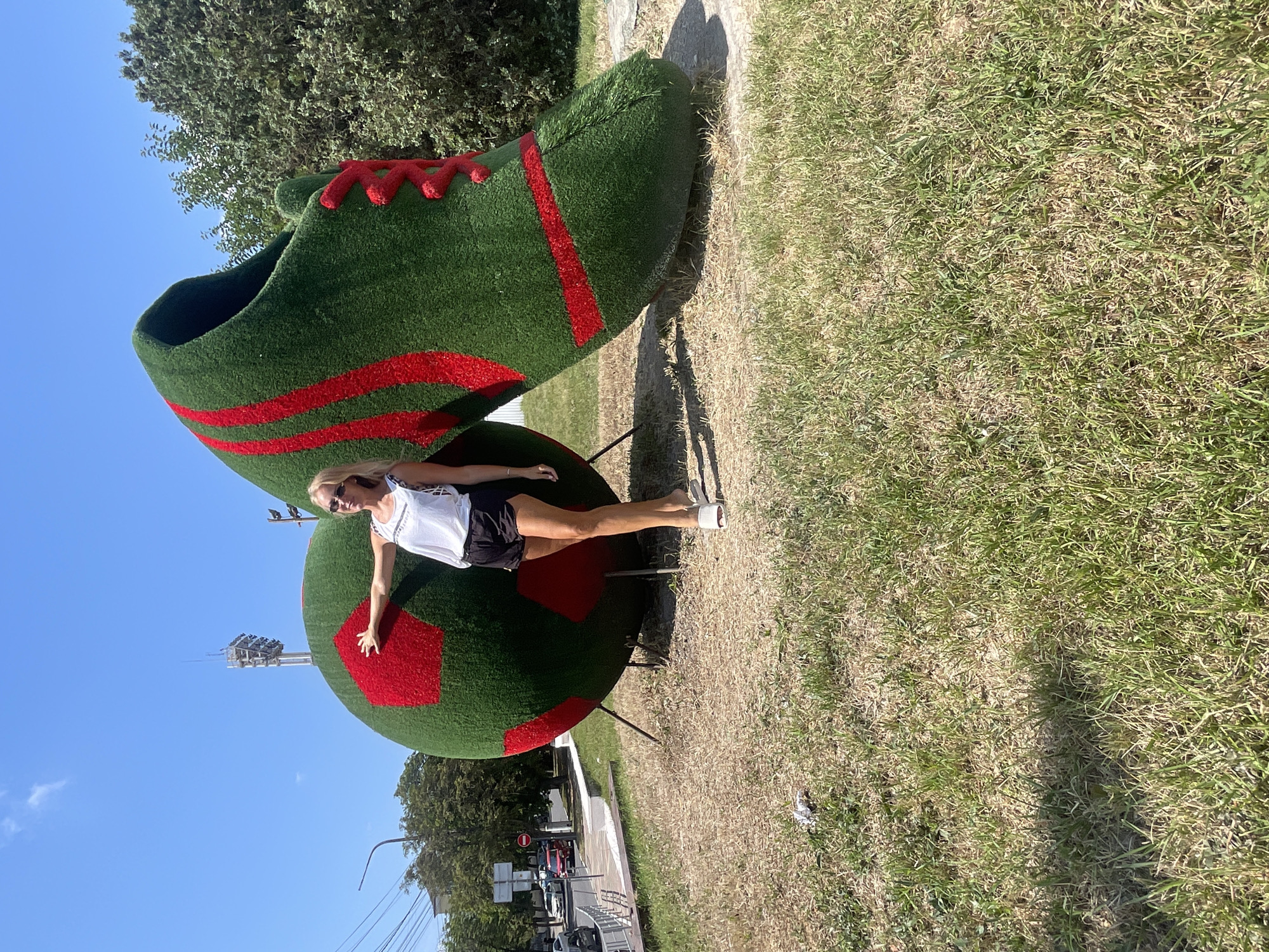 A woman poses in front of a large, green, soccer-themed sculpture in Novorossiysk, Russia. The sculpture depicts an oversized soccer ball with red pentagonal panels and a giant green cleat with red laces and stripes. The woman, wearing a white tank top, black shorts, and white sandals, stands between the ball and the cleat, with her left hand resting on the soccer ball. The sculpture is situated on a dry, grassy area with a slight incline. In the background, a street with cars and pedestrians is visible, along with trees and a stadium light tower. The sky is clear and blue.