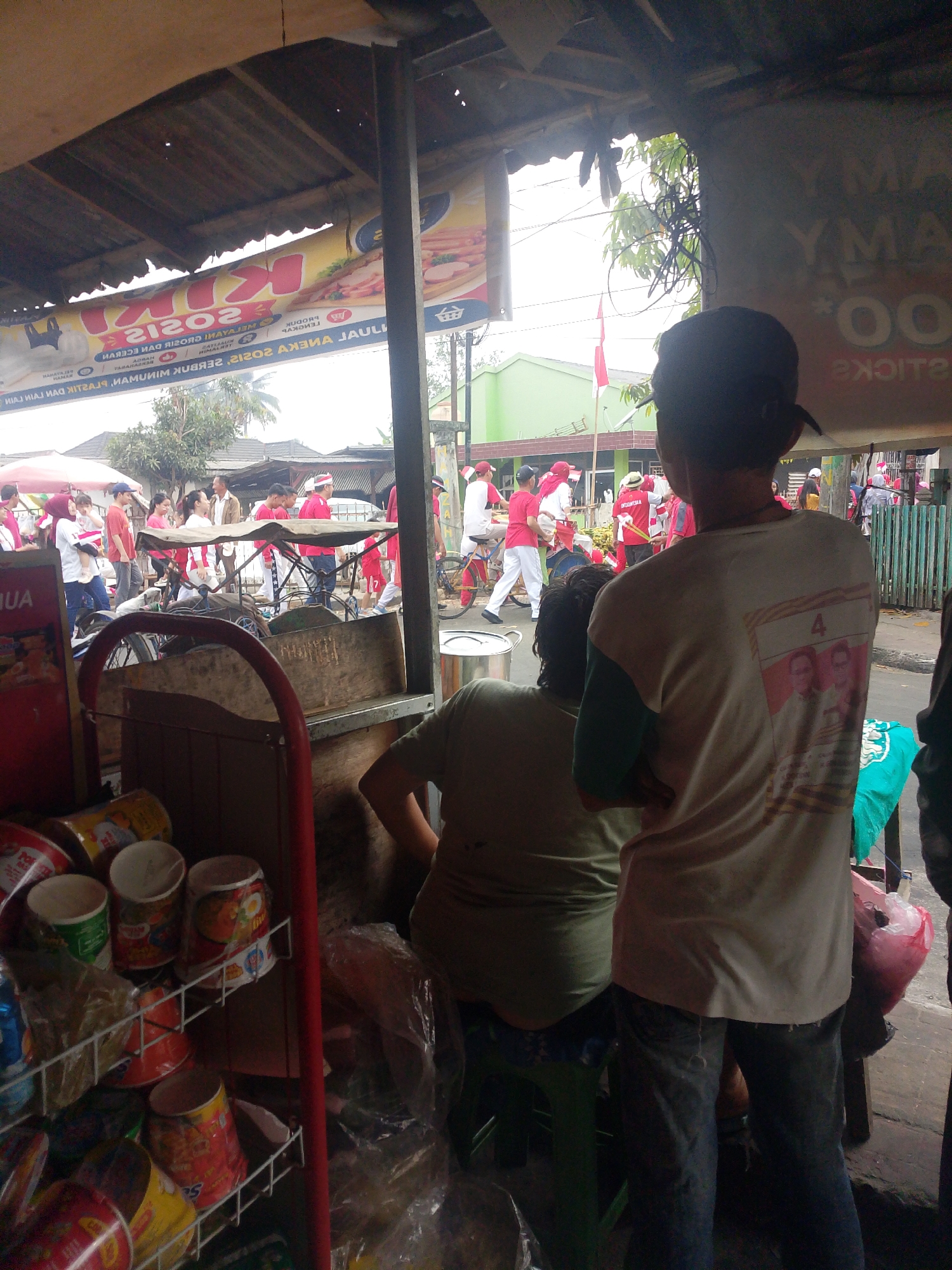The image captures a street scene in Palembang, Indonesia, likely during a daytime event or parade. In the foreground, two individuals are visible. The person on the right is wearing a light-colored t-shirt with a graphic of three faces on the back and dark pants. The person on the left is wearing a light green t-shirt and is seated.
Behind them, a marketplace or roadside stall is set up, with shelves displaying various food items in cans or cups, some wrapped in plastic. A red metal frame structure is prominent, supporting these shelves. In the mid-ground, a crowd of people, many wearing red and white attire, are gathered. Some are on bicycles, while others are walking. A red and white Indonesian flag is visible in the background.
The environment appears to be an urban setting with buildings and trees. The overall impression is one of a lively community gathering, possibly related to a local celebration or political event. The banner hanging overhead in the background, though partially obscured and with text in a foreign script, suggests commercial activity. The lighting indicates it is daytime.