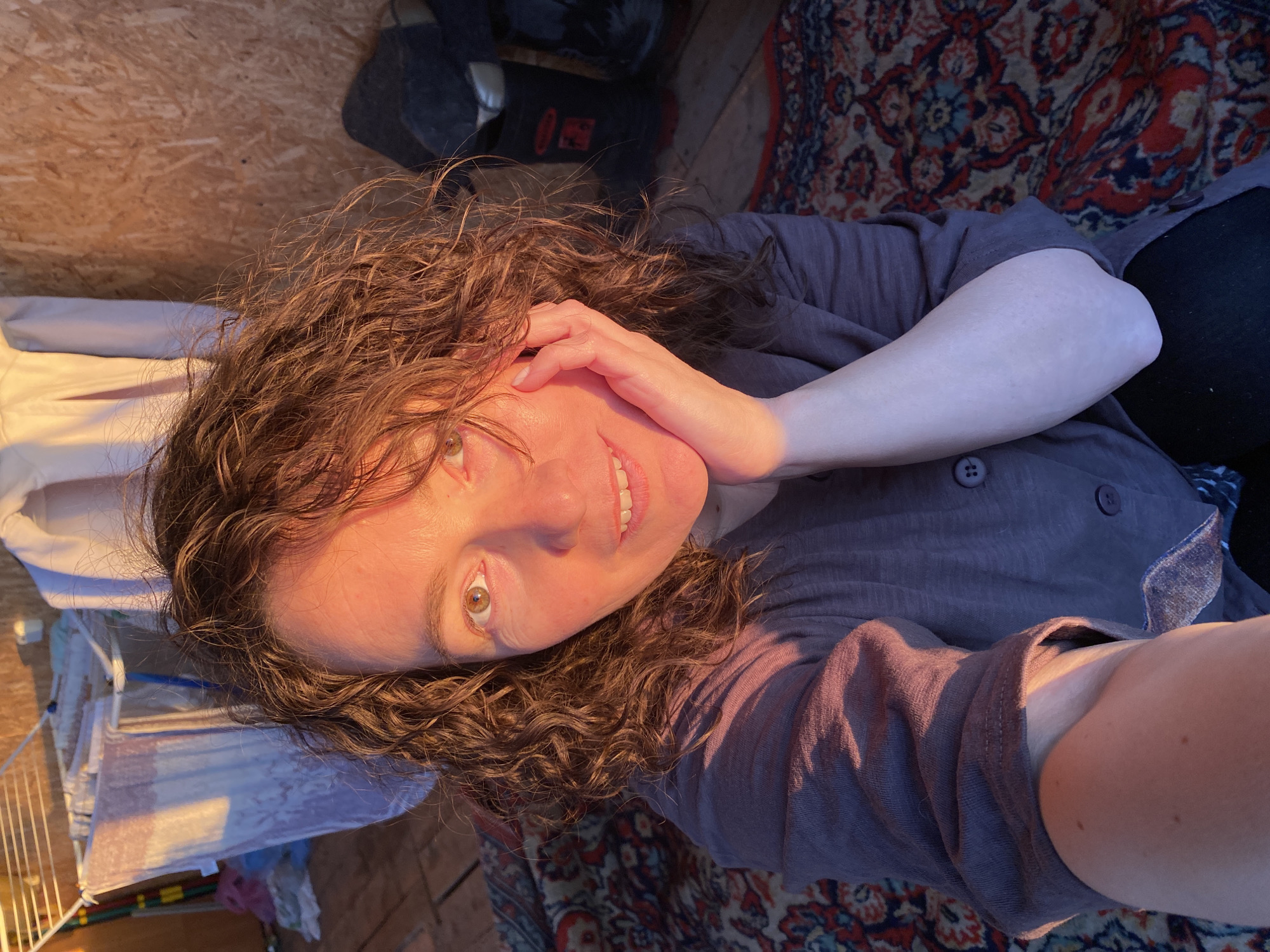 A woman with curly hair is lying on a patterned rug. She is wearing a long-sleeved grey shirt. Her right hand is near her face. Light illuminates one side of her face and hand. Behind her, a wooden wall is visible, and a drying rack with clothes is on the left side of the image. There is also an object that is either a container or a piece of luggage in the top corner. There is no evidence to confirm this image was taken in the city of Ольховка, Russia.