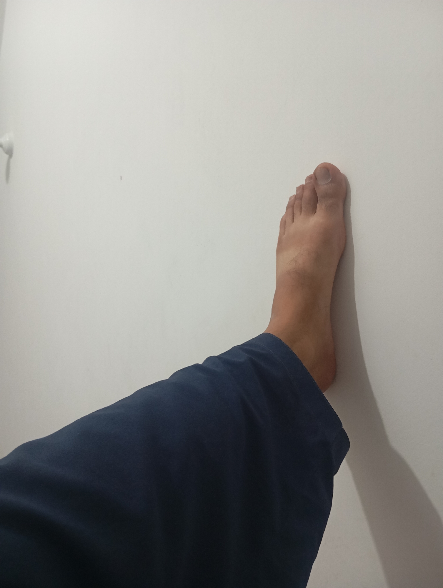 The image shows a person's bare foot and lower leg, clad in dark blue pants, resting against a plain white wall. The foot is positioned vertically against the wall, with the toes pointing upwards. A shadow cast by the foot and leg is visible on the wall to the right, indicating a light source from the left. There is a slight crease in the pants just above the ankle. The image is a close-up, focusing on the foot and lower leg against the wall. The environment appears to be indoors, with a simple, unadorned wall. The location in the city of Lassan, Pakistan is not determinable from the image itself.