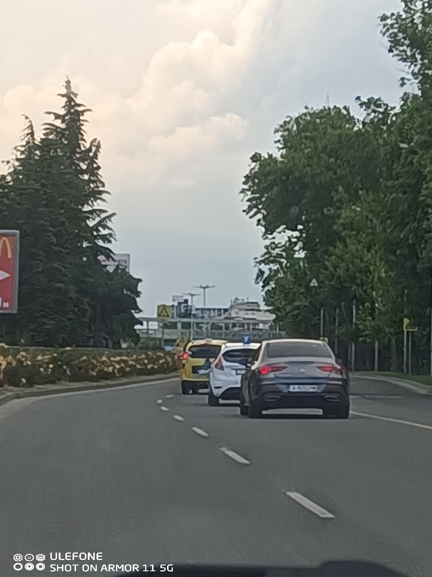 The image displays a forward-facing perspective from a moving vehicle on a multi-lane asphalt road, showing a line of three cars in traffic under an overcast sky. This location is identified as the approach road to Burgas Airport in Burgas, Bulgaria, specifically heading towards the main terminal building.
In the immediate foreground, the rear of a dark grey Mercedes-Benz CLA (C118 generation) is visible, featuring a Bulgarian license plate with the registration "A 820..." (partially obscured), confirming the Burgas region. Directly ahead of the Mercedes is a white Ford Fiesta. The third vehicle in the line is a yellow compact car, possibly a small van or taxi, positioned further down the road.
To the left, a landscaped median strip separates the road, planted with shrubs and light-colored flowering plants. Beyond this median, a large red and yellow McDonald's sign is partially visible behind tall, dark green conifer trees. Other advertising billboards are also discernible through the foliage.
To the right of the road, a dense line of deciduous trees runs parallel to the asphalt. A yellow triangular warning sign with a black exclamation mark is visible among these trees, and a fence can be partially seen behind the tree line. In the distant background, structures characteristic of an airport terminal building are present beneath a sky filled with large, light-colored clouds.
The road surface is asphalt, marked with white broken lines indicating traffic lanes. No individuals are explicitly visible outside of the vehicles. The image carries a "Ulefone Shot on Armor 11 5G" watermark in the lower left corner.