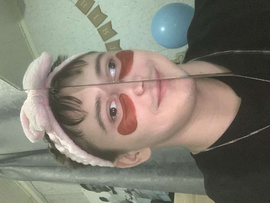 A youthful individual, likely male, is centered in a medium-shot portrait, wearing a pink, fluffy headband with a prominent bow and red gel under-eye patches. Their face is vertically bisected by a reflective surface, which creates a sharp division down the center of the frame. The individual has a faint mustache and a subtle smile, with their left eye gazing forward and their right eye directed slightly to the right. They are wearing a dark-colored t-shirt and a thin silver chain necklace. The background on the left consists of a grey curtain and a visible portion of a black and white image of a person in a suit. On the right, a light-colored wall is visible, adorned with a light blue spherical balloon and a banner displaying reversed Cyrillic letters, consistent with a celebratory decoration. The scene takes place indoors in a room in Foki, Russia.