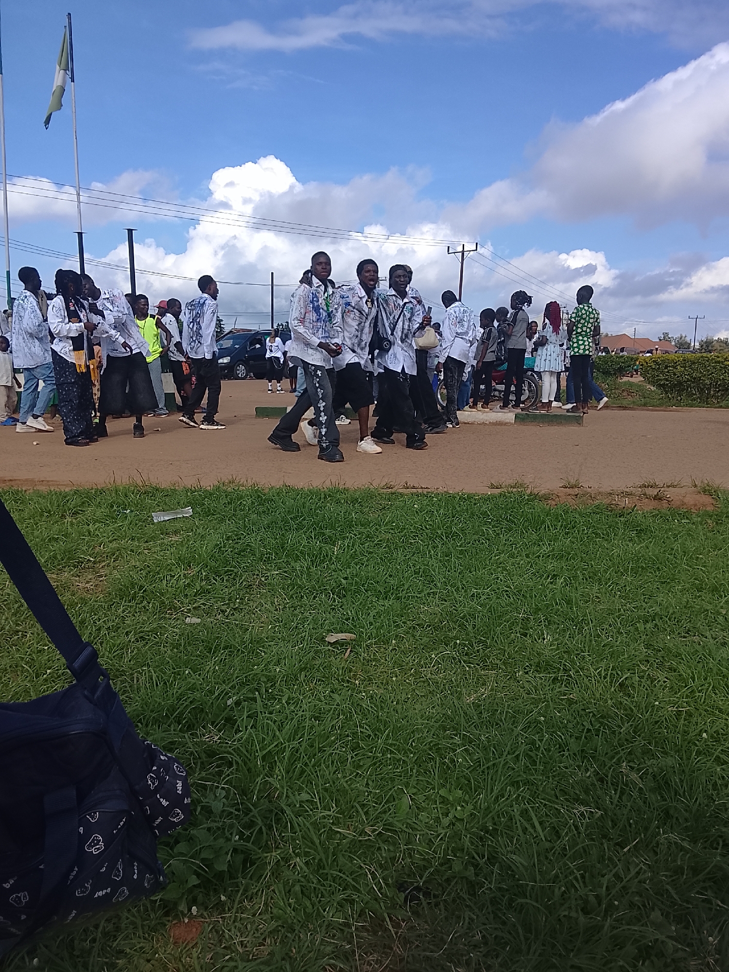 An outdoor scene depicts a group of people, primarily young men and women, gathered on a paved surface. The group is diverse in attire, with many wearing white shirts adorned with colorful drawings or patterns, suggesting a possible event or uniform. Some individuals are holding or carrying items, and a few are interacting amongst themselves. In the background, a flag on a pole is visible against a partly cloudy blue sky. Power lines and a dark-colored vehicle are also present. The foreground is dominated by a patch of green grass, with a dark blue bag with a pattern of white shapes lying on it. The scene appears to be in a semi-urban or campus environment.