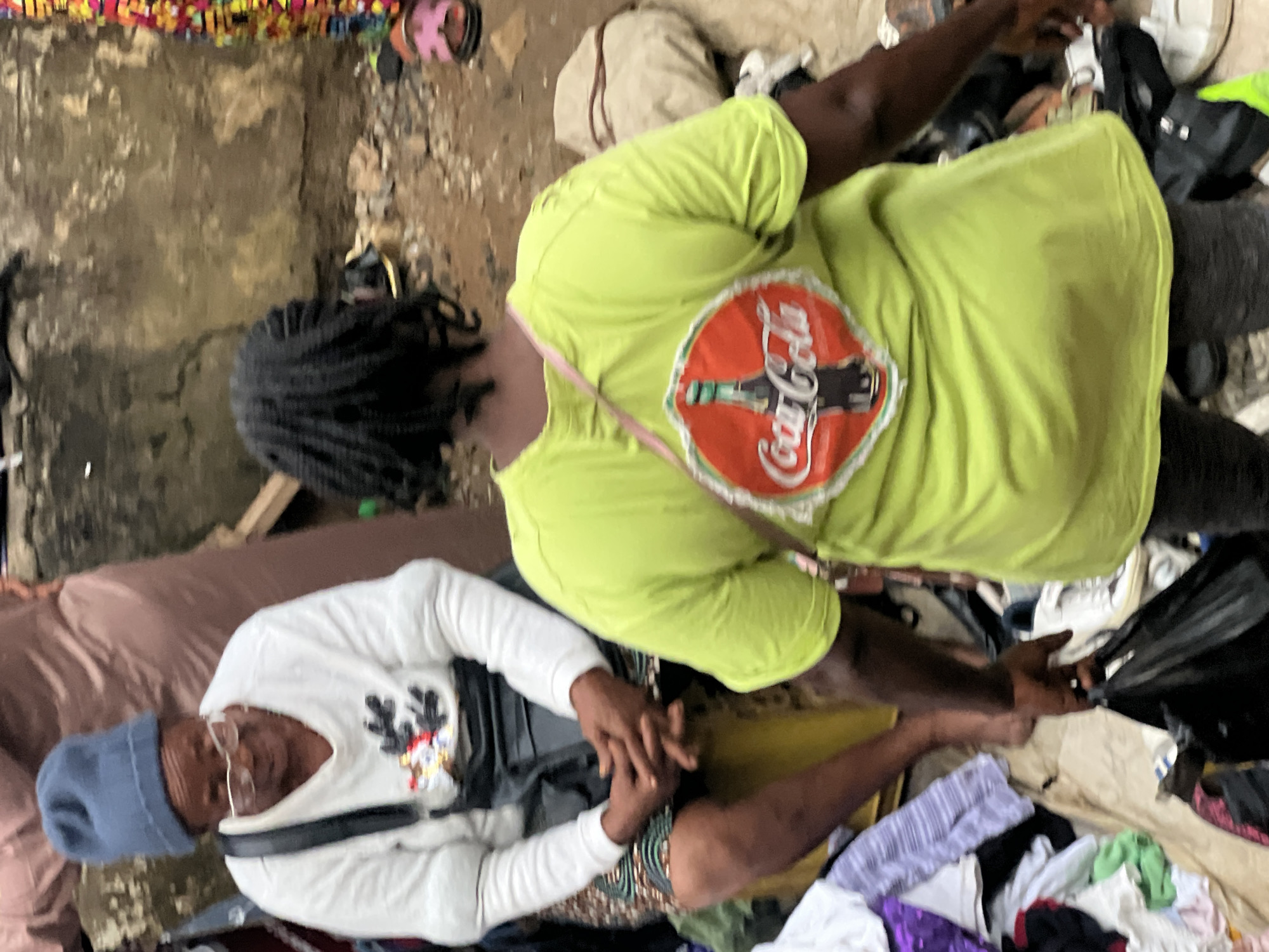 Two individuals are positioned amidst a collection of miscellaneous items. The person on the left wears a white long-sleeved shirt with a black belt, a blue knitted cap, and spectacles. The person on the right wears a lime-green short-sleeved shirt adorned with a Coca-Cola logo. They clasp each other's hands, the surroundings cluttered with clothing, bags, and other objects scattered about. The setting appears to be Idimu, Nigeria.