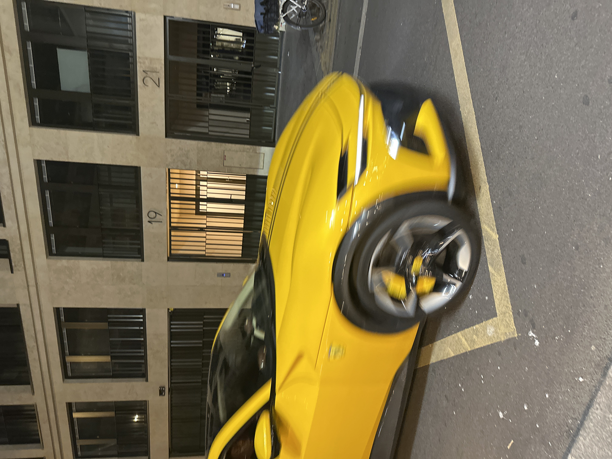 A blurry photo taken at night depicts a yellow Ferrari parked adjacent to a building, identified by prominent numerals 19 and 21, in what appears to be the city of Paris, France. A bicycle is parked nearby, but its details are indistinct. The photograph's focus is not sharp, resulting in a motion blur effect that obscures fine details of the car and the surrounding environment.