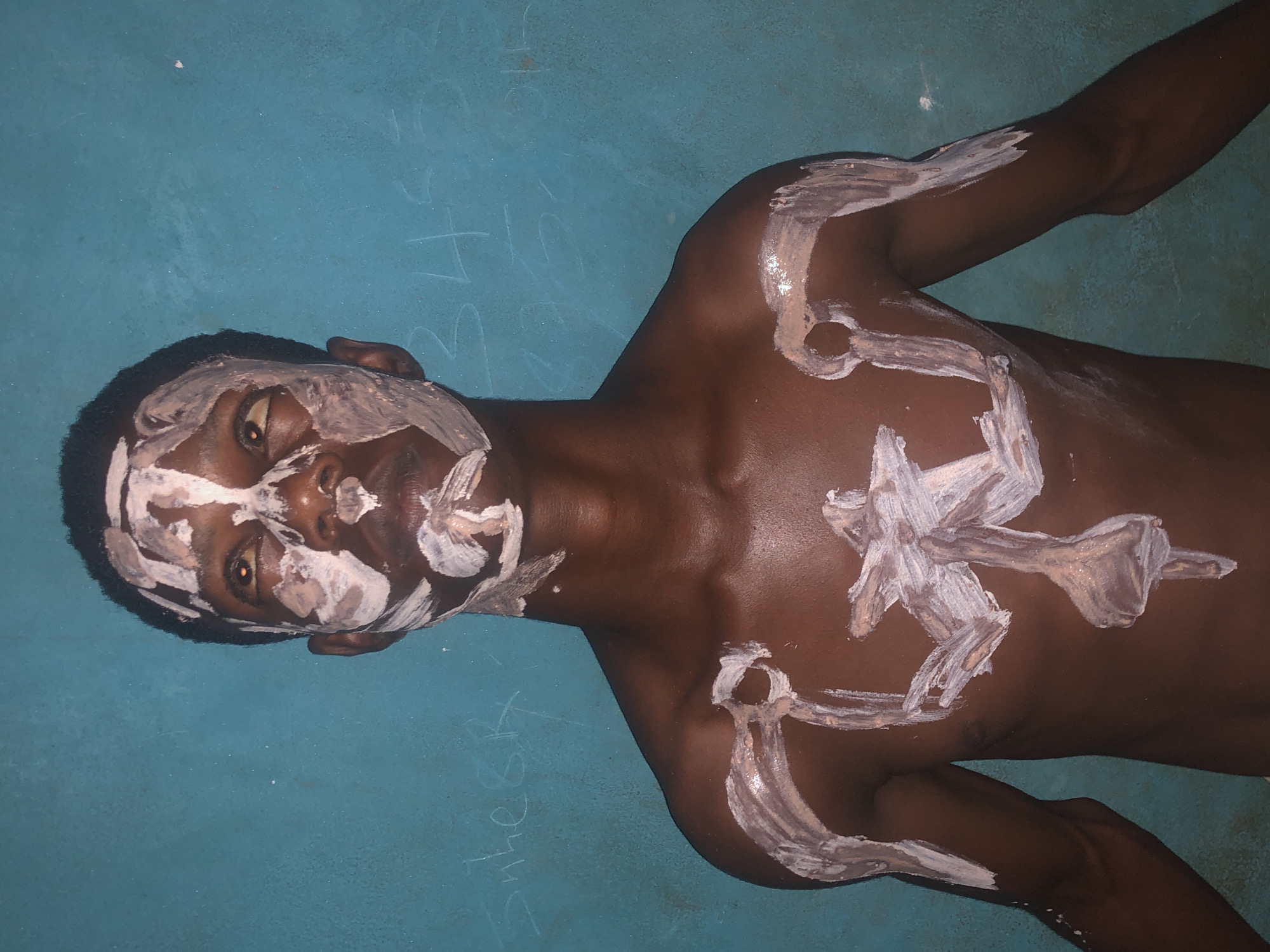 A shirtless, dark-skinned man with short hair is lying on a blue surface. He has white paint or cream smeared on his face and body in abstract patterns. The paint covers most of his face, as well as his chest, shoulders, and arms. In the lower left corner, there appears to be writing in chalk on the blue surface, possibly the word "Sulkeek." The background appears to be a flat, slightly textured blue surface, possibly a wall or floor. The image does not provide information about the location in the city of Bende, Nigeria.