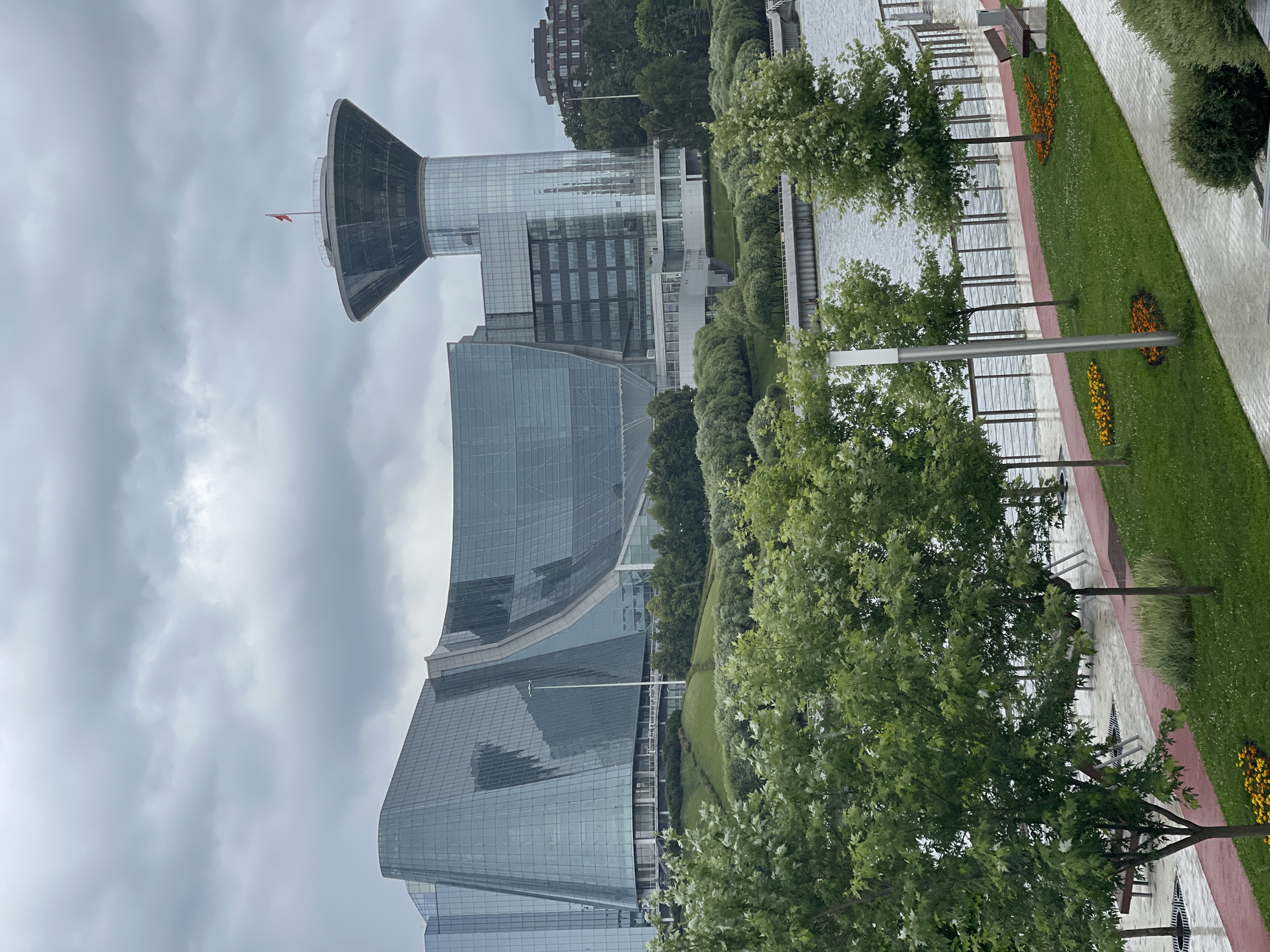 The image captures a modern urban scene on a cloudy day in Khimki, Russia. The main subjects are several contemporary glass-facade buildings, including one with a distinctive, wide, conical top structure. A red flag is visible on a pole extending from this conical section. The architecture is sleek and geometric, with reflective surfaces that mirror the overcast sky.
The setting is an outdoor public space adjacent to a body of water, likely a river or canal. A paved walkway runs along the water's edge, separated by a railing. Beyond the walkway, a neatly manicured green lawn is present, featuring decorative flower beds with yellow and orange blooms. Trees with lush green foliage are strategically placed throughout the park-like area, providing shade and natural beauty. Benches are visible along the walkway, suggesting a place for relaxation and enjoying the view.
The weather appears to be overcast, with a thick layer of clouds dominating the sky, contributing to a subdued lighting. No people are visible in the frame, giving the scene a sense of stillness. The overall impression is one of a well-designed urban waterfront, combining modern architecture with accessible green spaces. The time of day is indeterminate due to the cloud cover, but the diffused light suggests it is daytime.