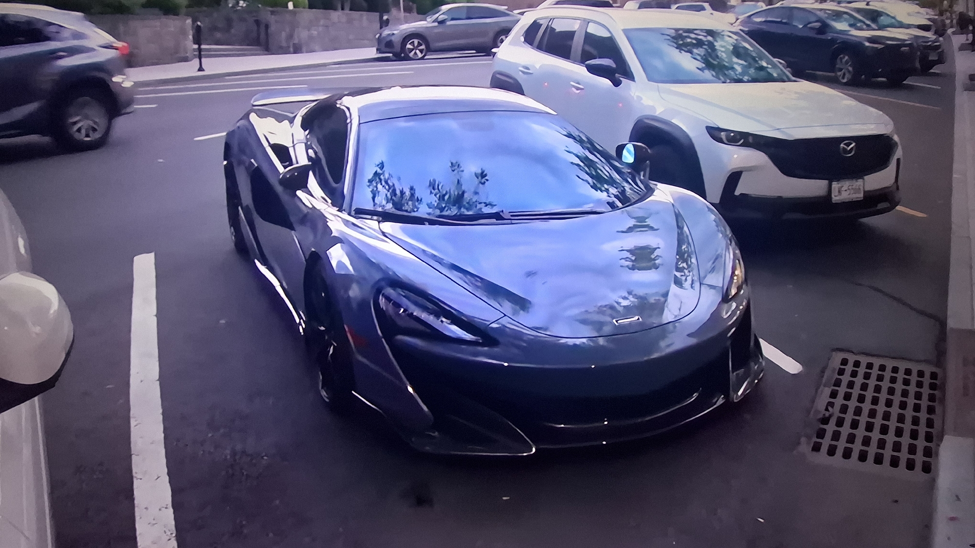 An eye-level shot captures a modern street scene featuring several vehicles. In the foreground and center, a dark blue or grey McLaren supercar is prominently parked, its sleek design and low profile evident. The windshield reflects trees and an overcast sky, suggesting it's daytime.
To the left of the McLaren, only a small portion of a light-colored vehicle, possibly white, is visible, showing part of its bumper and side mirror. Further left and slightly behind, a dark grey SUV (possibly a Lexus RX or similar model) is in motion, with only its rear-left side and wheel clearly visible.
To the right of the McLaren, a white Mazda CX-50 SUV is parked, facing the viewer. Its front grille and headlights are distinct, and a partial license plate is visible, reading "LK-5566". Behind the Mazda, another light grey sedan (possibly an Audi) is parked, and various other cars are visible further down the street in the background, including several SUVs and sedans.
The vehicles are situated on asphalt pavement with white painted parking lines. A concrete curb and a metal storm drain grate are visible in the lower right corner. In the background, there's a stone wall or building structure and some foliage, further suggesting an urban or suburban setting. The overall lighting indicates daytime, possibly an overcast day or late afternoon, with soft, diffused light. No people are visible in the image.
