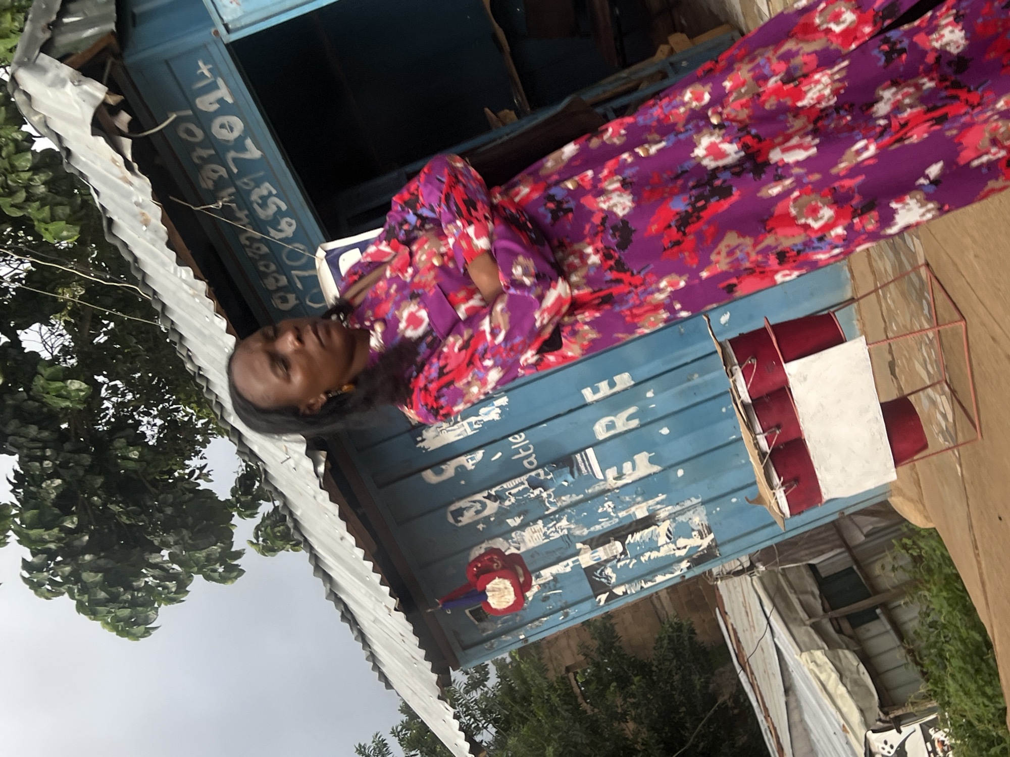 The image shows a woman in a vibrantly patterned purple, red, and white dress standing in front of a blue shipping container. The container appears to be a small shop or stall, with some peeling stickers and writing on its exterior. A corrugated metal roof extends over the front of the container, providing shade. To the left of the container, there is lush green foliage and a tree. To the right, a red metal frame structure, possibly a display stand, holds some items wrapped in red and white. The sky is overcast, suggesting a cloudy day. The woman is looking to her left, and her expression is neutral. The scene seems to be outdoors in a casual, possibly commercial, setting.
Visible text on the blue container includes what appears to be a phone number, "+234 8032859370" or similar, written in white lettering. Some other faded or partially obscured text is also present. The overall impression is a candid snapshot of daily life in Bukuru, Nigeria, capturing a woman near her place of business.