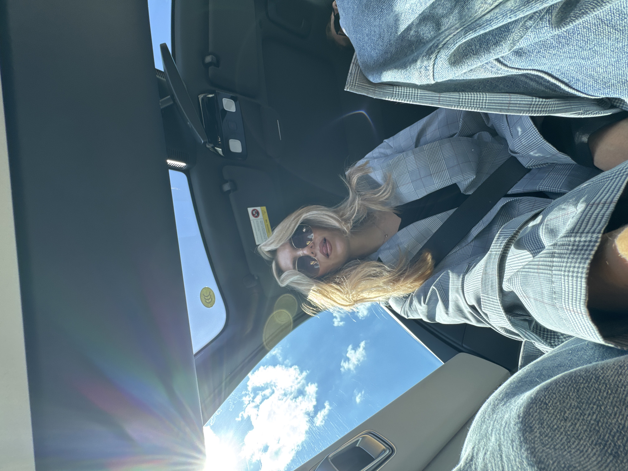 The image shows a blonde woman with sunglasses inside a car, wearing a gray and white checkered blazer with a black shirt underneath. She is wearing a seatbelt. The image is taken from a low angle, looking up through the car's sunroof. The sky is visible through the sunroof, with white clouds and a bright sun. The car's interior is visible, including the dashboard and steering wheel.
The location is unknown.