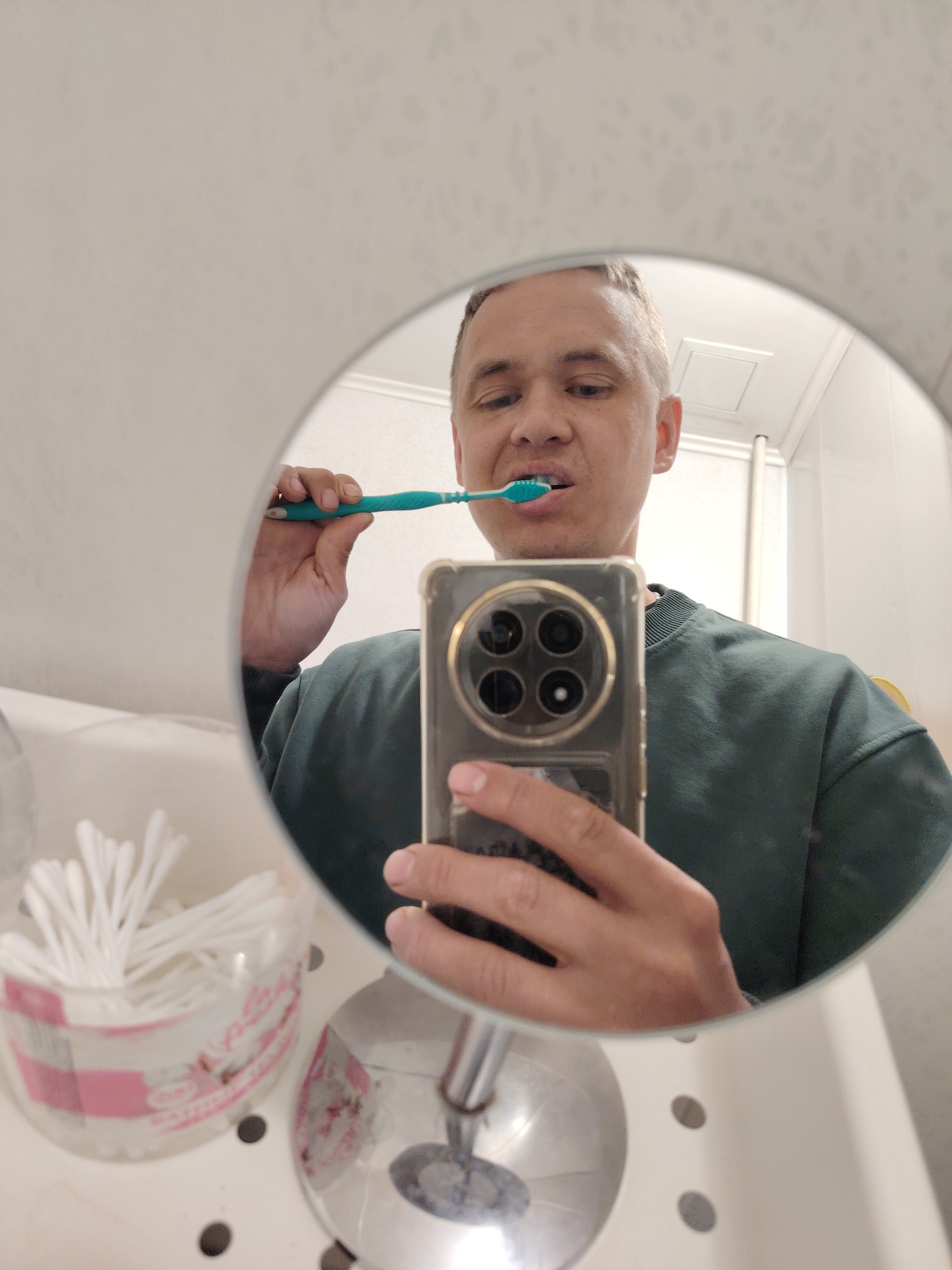 The image is a mirrored selfie of a man brushing his teeth in a bathroom. The man, with short light hair and wearing a dark green long-sleeved shirt, is captured in his reflection. He holds a teal-colored toothbrush to his mouth with his right hand, actively engaged in brushing. His expression is neutral and focused on the task.
He is taking the photo using a modern smartphone, held in his left hand, which is visible in the bottom-center of the frame. The phone has a distinct, large, circular camera module with multiple lenses. The circular mirror frames the man's upper body and head.
In the background of the reflection, parts of the bathroom are visible, including a white ceiling with a rectangular vent or light fixture, and a white wall or shower partition. To the lower left, on a white countertop or sink edge, sits a clear plastic container filled with white cotton swabs. The container has a pink label with text, partially legible, appearing to be in a non-English language (likely Russian, given the location context of Chaikovsky, Russia) and seems to include words like "ВАТНЫЕ" (VATNYE), indicating cotton swabs. The countertop also has several dark, circular indentations. The scene depicts a routine moment of personal hygiene, likely in the morning or evening.