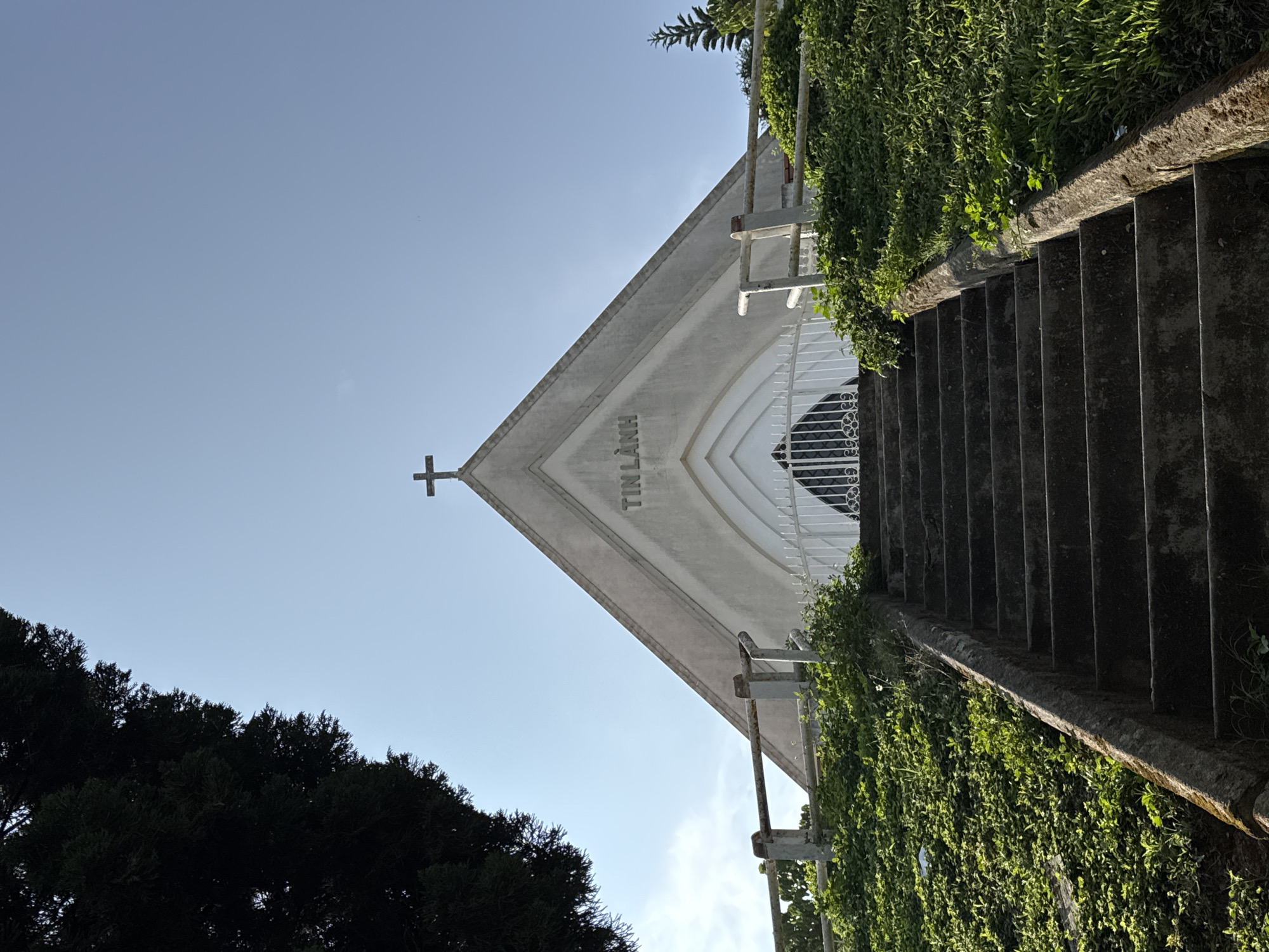 The image captures an upward view of a white, A-frame church, presumably a Protestant or Evangelical church given the visible text, located in Da Lat, Vietnam. The perspective is from the bottom of a wide, concrete staircase, looking up towards the building's facade and the bright sky.
The church features a simple cross at its apex, and the Vietnamese text "TIN LÀNH" is prominently displayed on the front gable. Below this inscription, an arched entrance or window area is adorned with decorative white metalwork. To the right of the church and along the ascending staircase, dense, vibrant green foliage and vines cover an adjacent wall or slope. Weathered metal railings run alongside the stairs, providing a handhold. On the left side, the dark, silhouetted crowns of trees contrast sharply with the clear, pale blue sky.
The scene is brightly lit by natural sunlight, indicating a clear day, likely during the morning or afternoon. There are no people visible, and no specific activity is taking place; the image focuses on the architecture and its natural surroundings. The overall impression is one of tranquility and architectural grandeur, set against a serene backdrop.