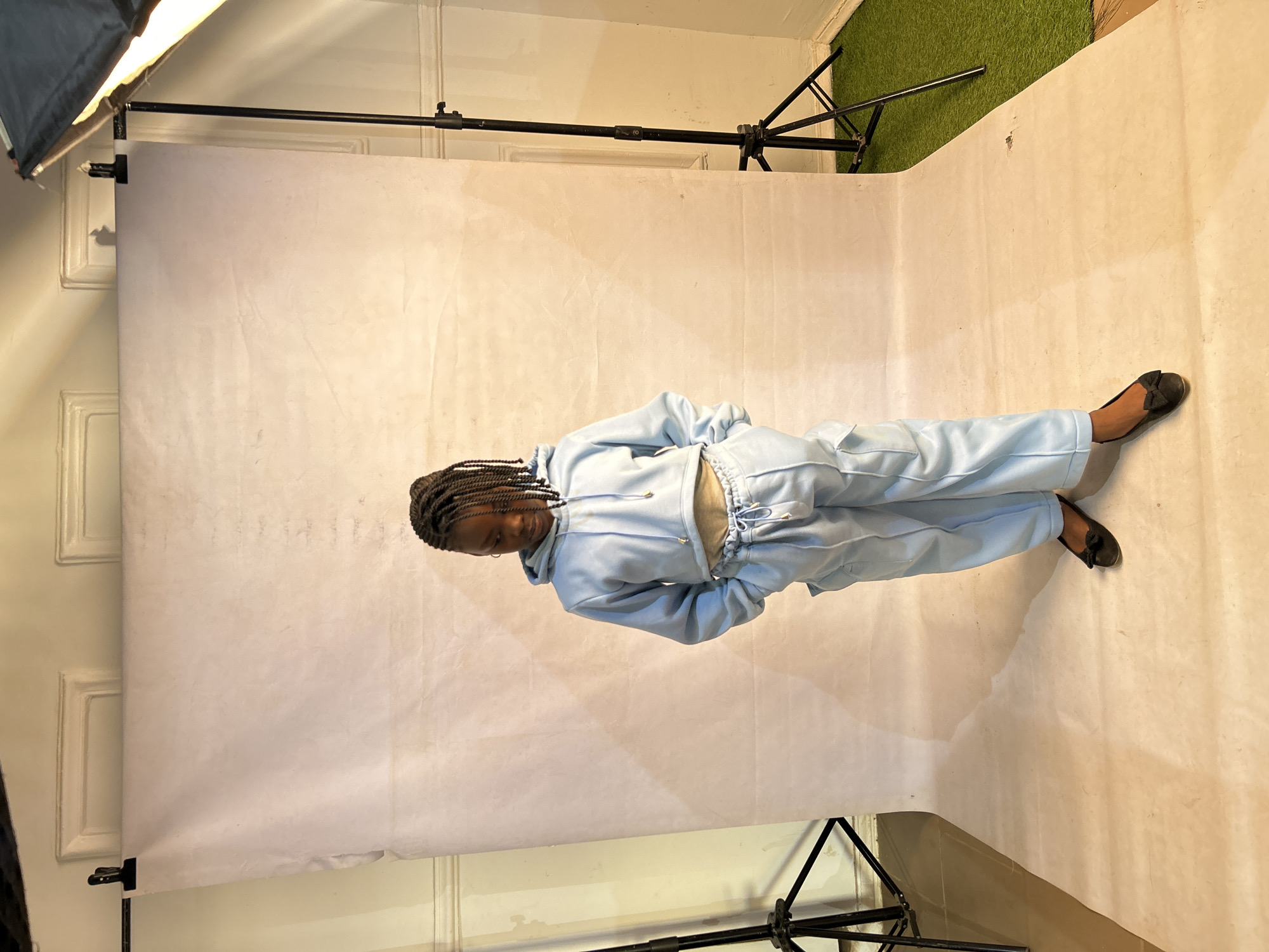 A person wearing a light blue tracksuit and black flats is lying on a white backdrop. The backdrop is supported by a stand with a softbox lamp attached to the top left. The backdrop is set up in a room with white walls and green turf at the top right corner. The location in Jos, Nigeria, is not verifiable from the image.