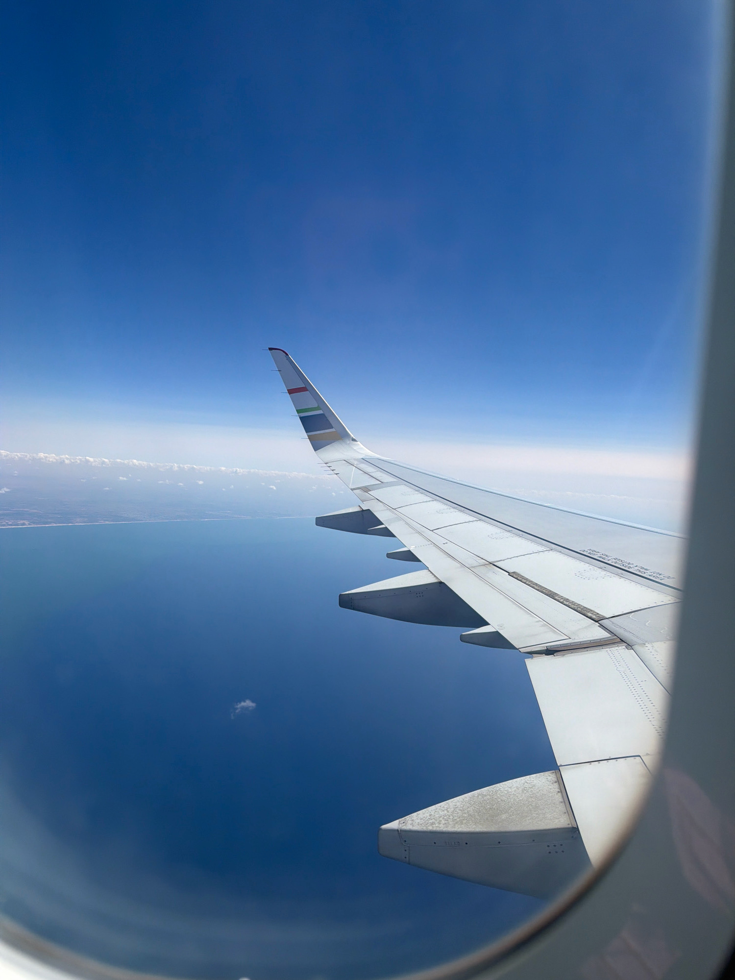 This is an aerial view from an airplane window looking out over the Mediterranean Sea towards the coast of el Prat de Llobregat, Spain. The image features a portion of the airplane's wing extending into the frame from the right. The wing is white with visible rivets and panel lines. The wingtip is decorated with horizontal colored stripes. Below the wing, the deep blue of the sea is visible, meeting a hazy shoreline with a thin line of clouds above it. The sky is a clear, bright blue. The scene appears to be captured during daylight hours, suggesting good visibility.