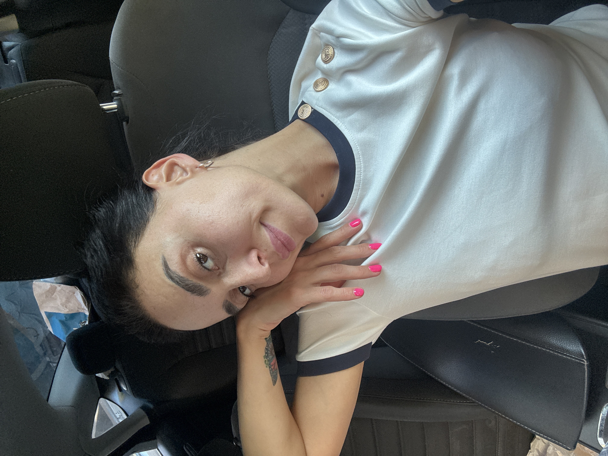 A woman with dark hair and fair skin is lying on her side, propped up against a car seat. She is wearing a white shirt with navy trim and gold buttons. Her left hand is resting on her cheek, and her right hand is on her chest. Her nails are painted bright pink. She has a small tattoo of a butterfly on her left forearm. Her expression is relaxed and serene. The background appears to be the interior of a car, with dark upholstery visible. The lighting suggests it is daytime. The photo was taken in Tbilisi, Georgia. There is no visible text in the image.