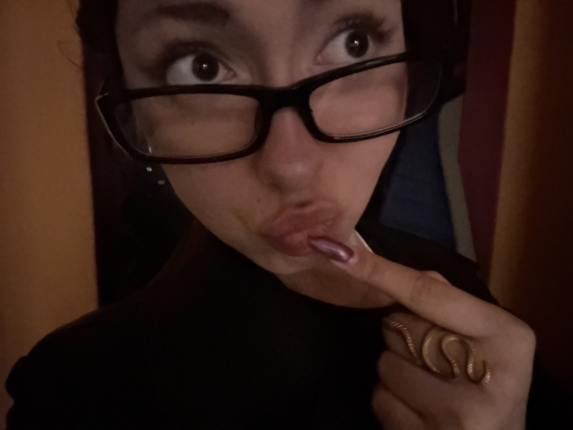 A person with brown eyes, wearing black-framed glasses, is looking upwards and to the right. The person has pursed lips and is touching their lower lip with the index finger of their right hand, which is adorned with a gold-colored ring shaped like a serpent. The person is wearing a black top. The background includes indistinct vertical stripes of orange, red, cream, and blue. The location cannot be determined from the information in the photograph.