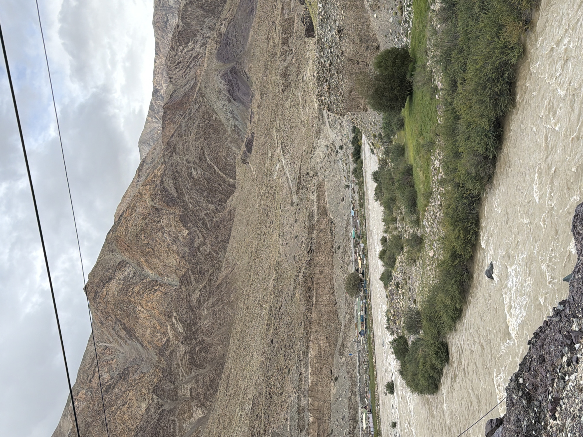 A muddy river flows through a valley with arid, rocky mountains. Several small buildings and tents are visible on the riverbank in the middle distance. A power line stretches across the sky. The weather appears overcast.