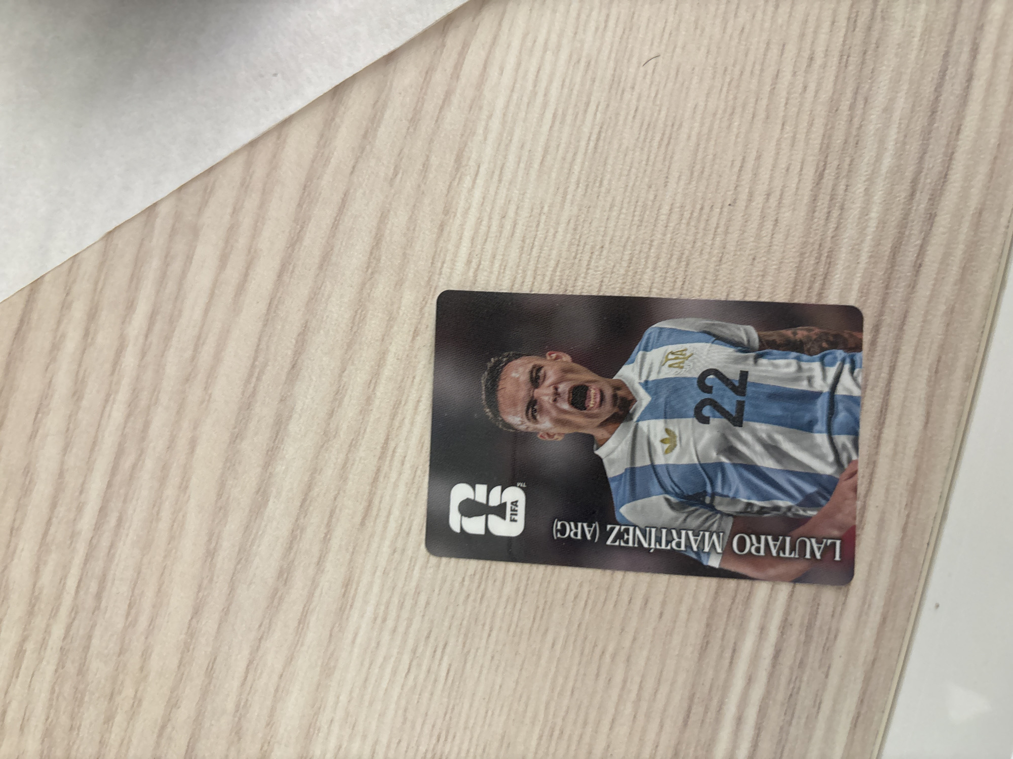 An image captured in Blagoevgrad, Bulgaria, displays a football trading card placed diagonally on a light-colored wooden surface. The card features a close-up portrait of Argentine footballer Lautaro Martinez, captured in a moment of intense emotion, possibly celebration or shouting, with his mouth wide open. He is wearing the light blue and white striped jersey of the Argentina national team, with the number "22" prominently displayed on the front. A small Adidas logo is visible on his right shoulder.
The card itself is dark-bordered, with a white, stylized "S" logo and the text "FIFA" stacked vertically in the bottom left corner (when viewed with the player upright). The player's name, "LAUTARO MARTINEZ (ARG)", is printed upside down along the bottom edge of the card. The wooden surface has a distinct grain pattern, running diagonally from the top left to the bottom right. In the upper left corner of the frame, a portion of a white or light grey object or surface is visible, contrasting with the wood. The overall setting appears to be indoors, likely on a desk or table.