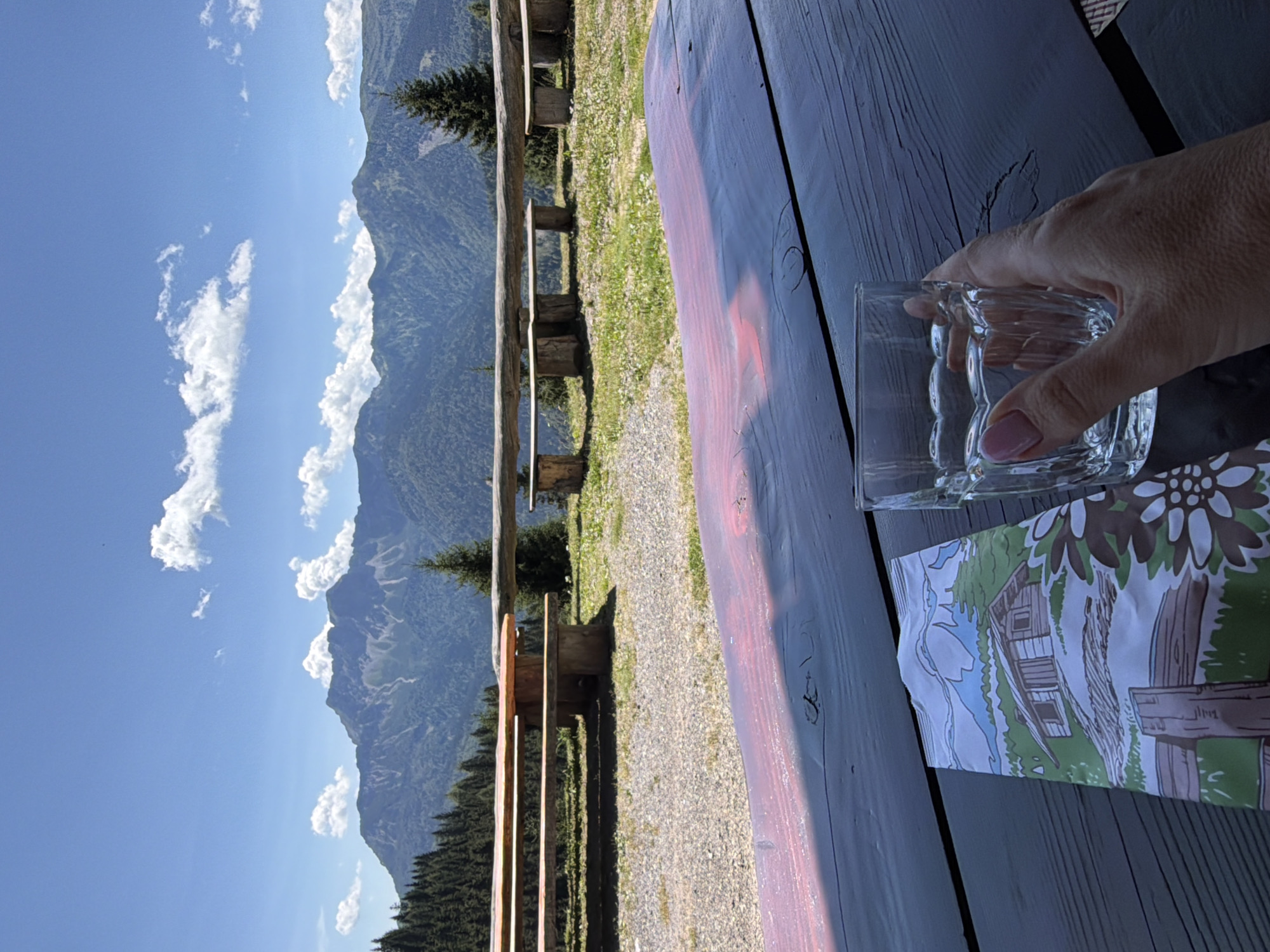 The image depicts a scenic outdoor setting, likely a mountain vista. In the foreground, a dark wooden table is visible, with a patterned brochure and a glass of water held by a person's hand. The brochure features an illustration of a rustic cabin amidst a green landscape with mountains in the background. Beyond the table, a gravel area leads to a wooden bench and picnic table arrangement, constructed from logs and planks. Further in the distance, a dense forest of evergreen trees covers the slopes of rolling mountains, culminating in a majestic, layered mountain range under a bright blue sky with scattered white clouds. There is no visual information in the image to identify the location as Lateis/Latais, Italy.