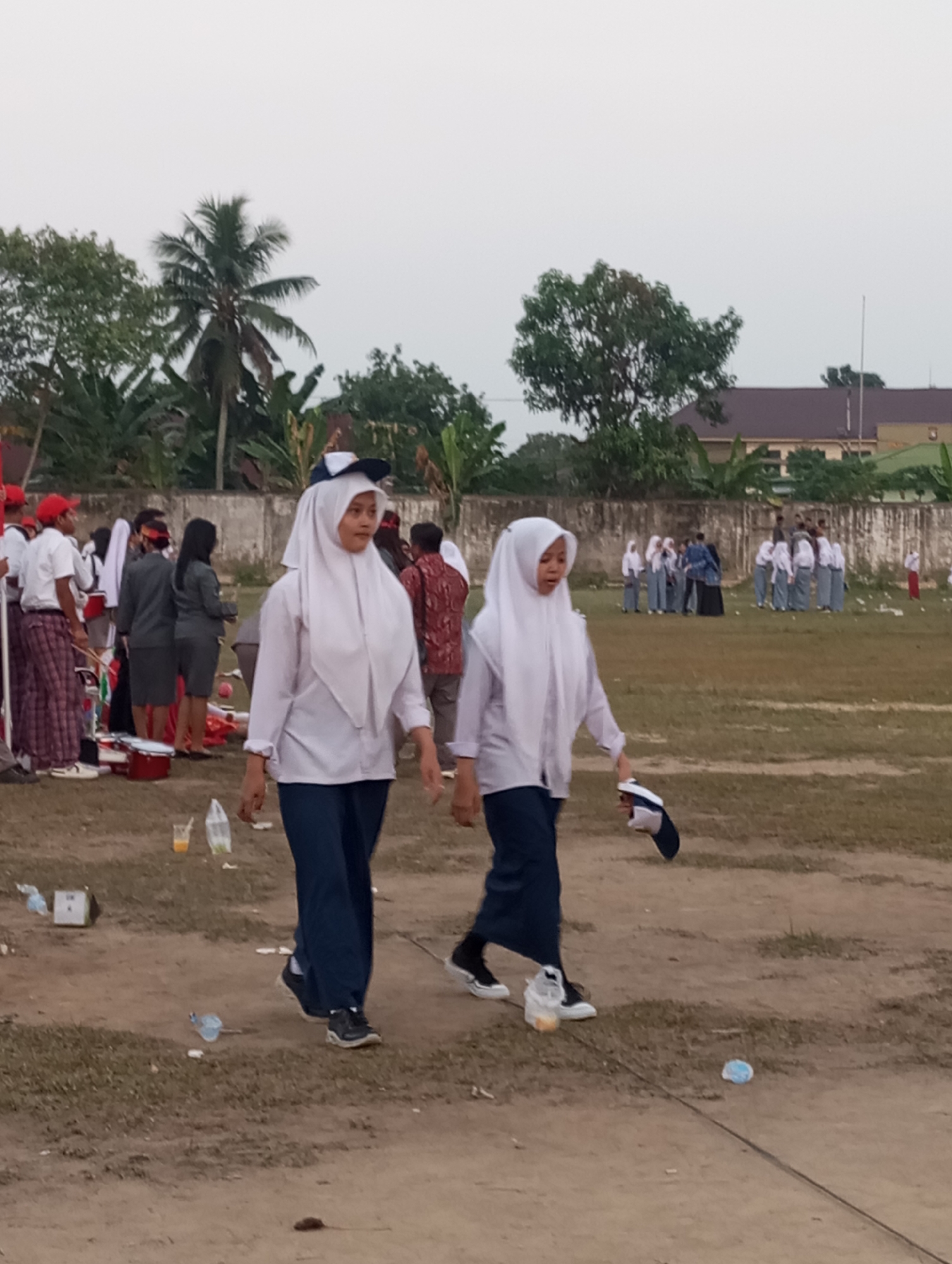 Two young women in white headscarves and blue pants walk across a dirt and grass field in Candirejo, Indonesia. The woman in the foreground on the left is wearing a white long-sleeved shirt and a white headscarf, and her dark blue pants are visible. She is looking to her right. She is holding a dark blue and white cap in her right hand. The woman to her right is also wearing a white long-sleeved shirt and a white headscarf, with dark blue pants. She is looking to her left and is slightly behind the first woman. She is wearing white sneakers with black laces.
In the background, several other people are visible. To the left, a group of people in red caps and white shirts, some with patterned red and white pants, stand near red cylindrical objects. Behind them, a woman in a grey uniform and another in a black headscarf and grey uniform are visible. Further in the background, a larger group of individuals, many wearing white shirts and grey pants, are gathered. A beige-colored building with a dark roof is visible in the far background, with trees and palm trees surrounding it. The ground is a mixture of dry dirt and patches of grass, with scattered litter including plastic bottles and cups. The sky is overcast.