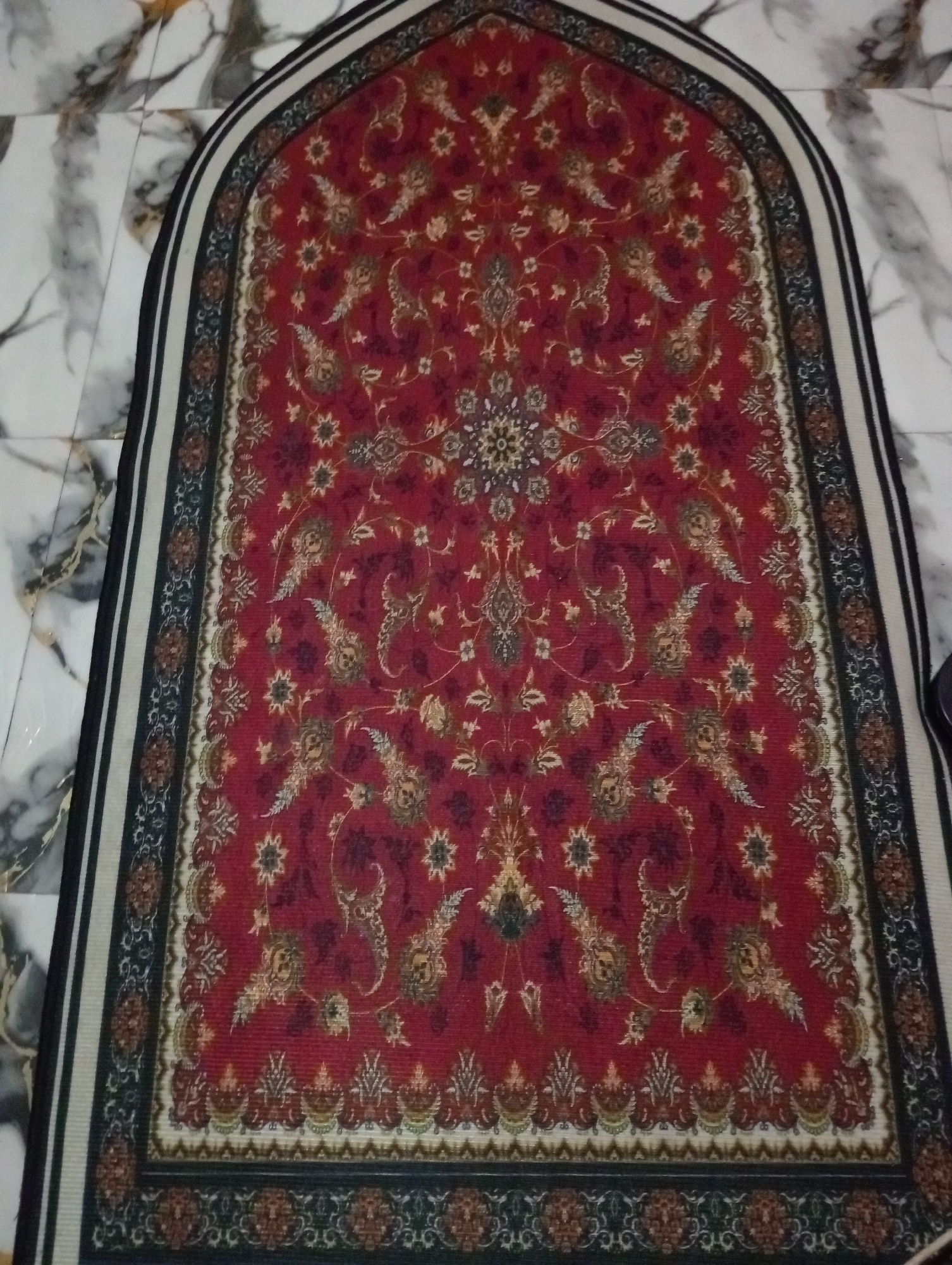 A red prayer rug with intricate gold and dark blue floral patterns is laid out on a marbled floor. The rug has a pointed arch shape at the top and a detailed border. The floor tiles are white with gray veins. The image appears to be taken from a high angle, looking down at the rug. No people are visible in the image. The setting is likely a room in a home or a place of worship. The lighting suggests it is indoors, possibly during the day with ambient light.