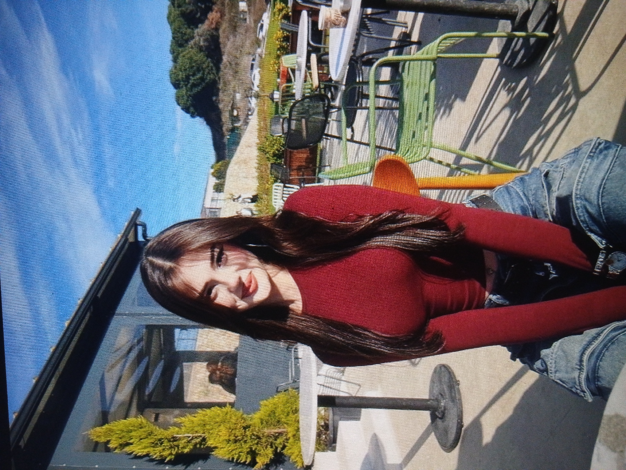 A young woman with long, dark hair is seated outdoors, facing the camera. She is wearing a maroon long-sleeved top and denim pants. The background suggests an outdoor seating area of a cafe or restaurant, with tables and chairs visible. There is a building with large windows and a tree to her left. Beyond the seating area, a grassy slope with trees and some parked cars can be seen under a bright blue sky with scattered clouds. The image quality is pixelated, and the lighting suggests it was taken during the day. No specific landmarks of Dnipro, Ukraine are discernible.