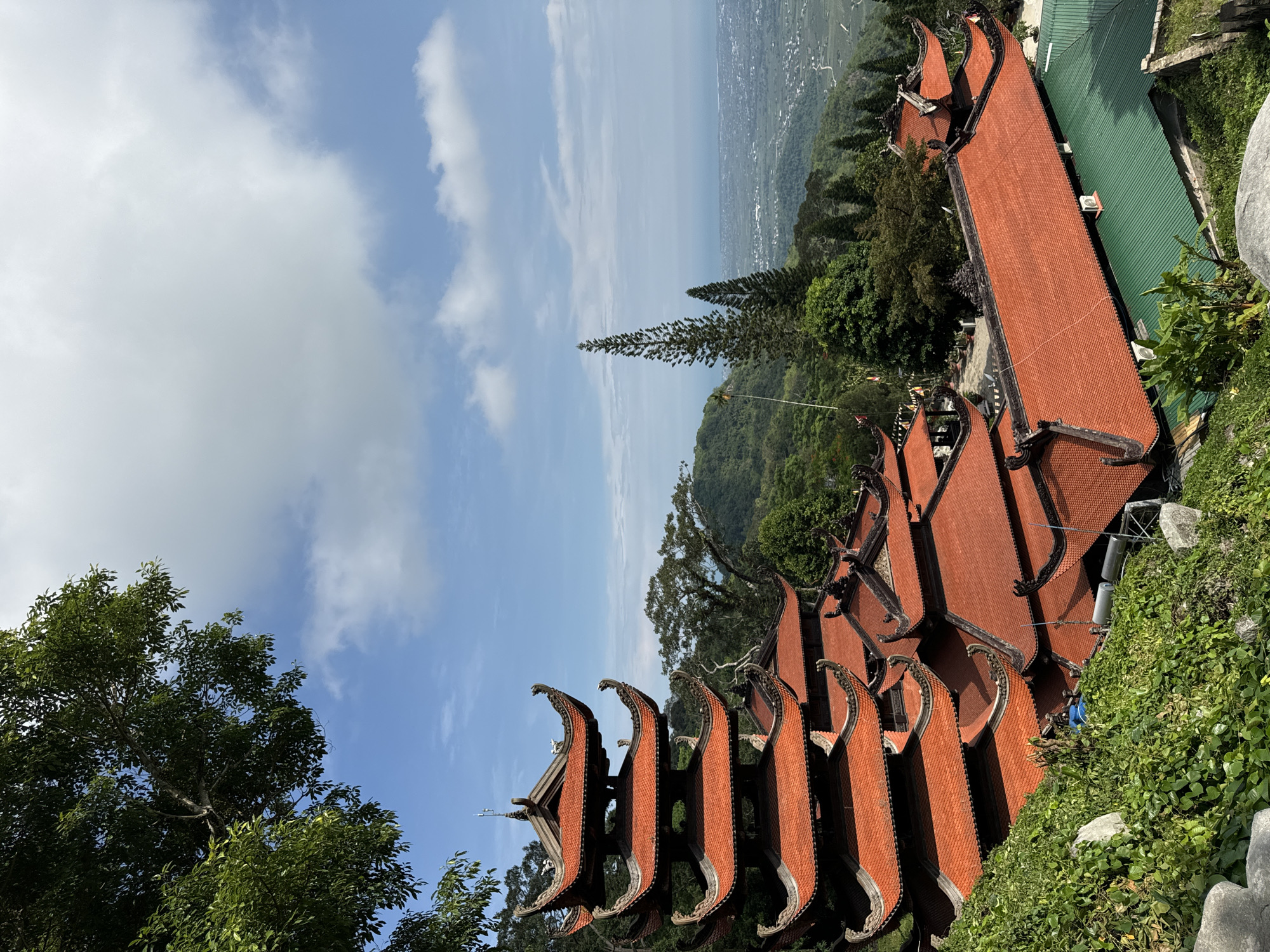 The image captures a panoramic view of a temple complex with distinct, ornate red-tiled roofs, set against a lush, mountainous landscape. The traditional architecture, characterized by curved eaves and detailed embellishments, suggests a location in Southeast Asia, consistent with the provided context of Thuận Nam, Vietnam. The temple appears to be situated on a hillside, offering a sweeping vista of a town or village nestled in the valley below, which stretches towards a distant body of water.
The sky above is a bright blue, punctuated by fluffy white clouds, indicating fair weather and likely daytime. The sunlight casts shadows, highlighting the textures of the tiled roofs and the dense green foliage surrounding the structures. A tall, slender evergreen tree stands prominently on the slope, its unique shape adding to the visual interest of the scene.
The image is taken from a high vantage point, looking down and across the temple buildings and the expansive landscape. There are no visible people in the frame, nor any explicit signs of an ongoing event. The focus is on the serene beauty of the temple architecture and its harmonious integration with the natural environment. The overall mood conveyed is one of peace and grandeur, emphasizing the spiritual and aesthetic qualities of the location. The presence of modern elements like air conditioning units attached to the temple walls provides a subtle contrast to the traditional design.