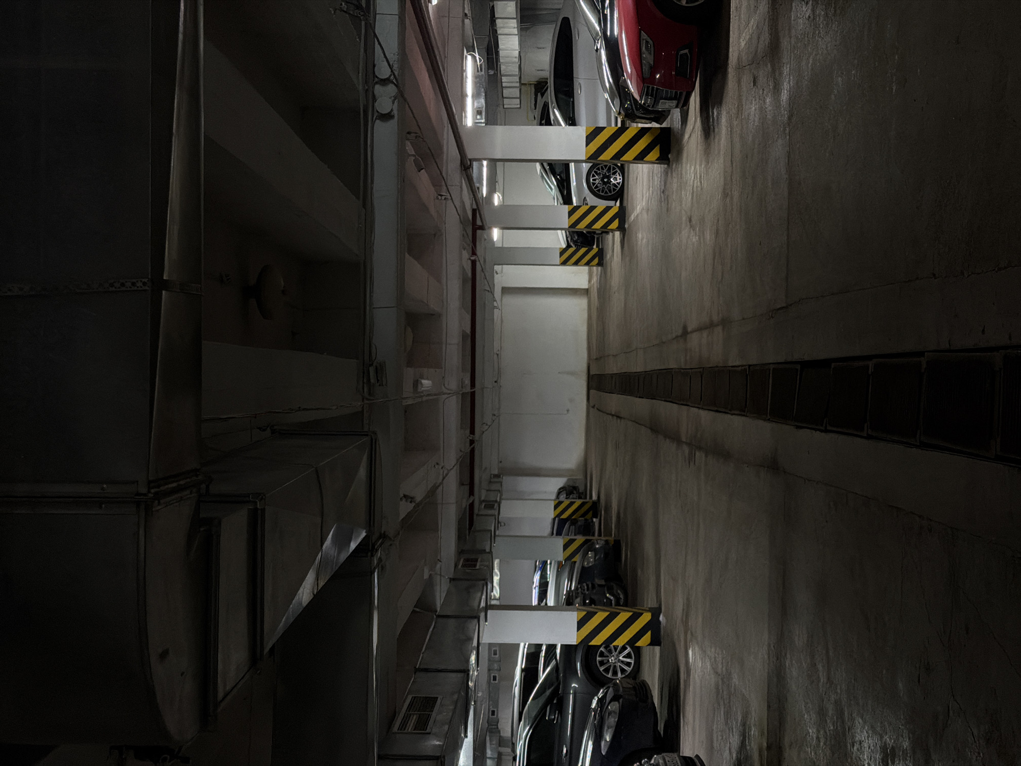 This image captures a stark, vertically-oriented view into what appears to be a multi-level parking garage or an industrial building in Krasnoyarsk, Russia. The perspective looks down a deep, narrow shaft or void, with structural elements and vehicles arranged vertically.
On the right side of the image, several cars are parked, facing into this central void. They are situated on different levels, appearing stacked one above the other due to the photograph's vertical orientation. The most prominent vehicles include a red sedan at the top, followed by a silver car, and several darker-colored passenger cars further down. Each parking spot is delineated by distinctive yellow and black striped barriers, which serve as car stops.
The left side of the shaft features a complex system of large, dark metal ducts and ventilation pipes running along the concrete wall. Exposed fluorescent light fixtures are visible on the walls, casting a dim, artificial light throughout the space. The concrete surfaces of the walls and structural beams show a utilitarian finish, consistent with industrial or parking facilities.
No people are visible, and the scene is static, depicting only parked cars and the building's infrastructure. The artificial lighting prevents determination of the time of day. The overall atmosphere is enclosed and industrial. No discernible text or specific location cues beyond the architectural elements are visible.