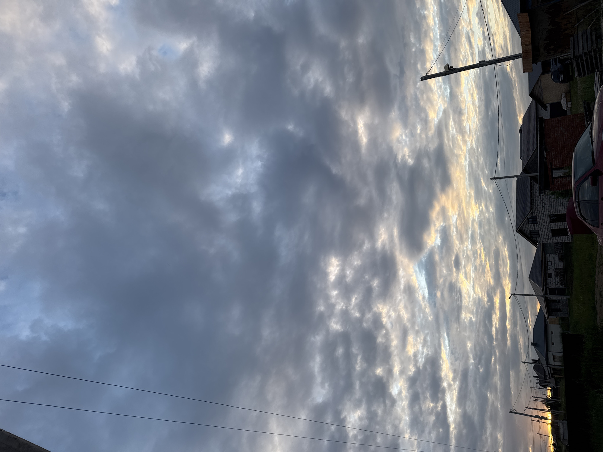 This is a vertical, eye-level, full shot of a cloudy sky over a residential area in Chaikovsky, Russia. The sky is filled with a variety of clouds, ranging from dark gray to light white, with patches of yellow and orange light peeking through, suggesting either sunrise or sunset. In the foreground, several houses with pitched roofs are visible, some constructed of brick and others with lighter colored siding. Power lines stretch across the frame, supported by utility poles. A red car is parked in the lower right corner of the image. The environment appears to be a suburban or rural setting, with grass and some scattered vegetation visible in front of the houses.