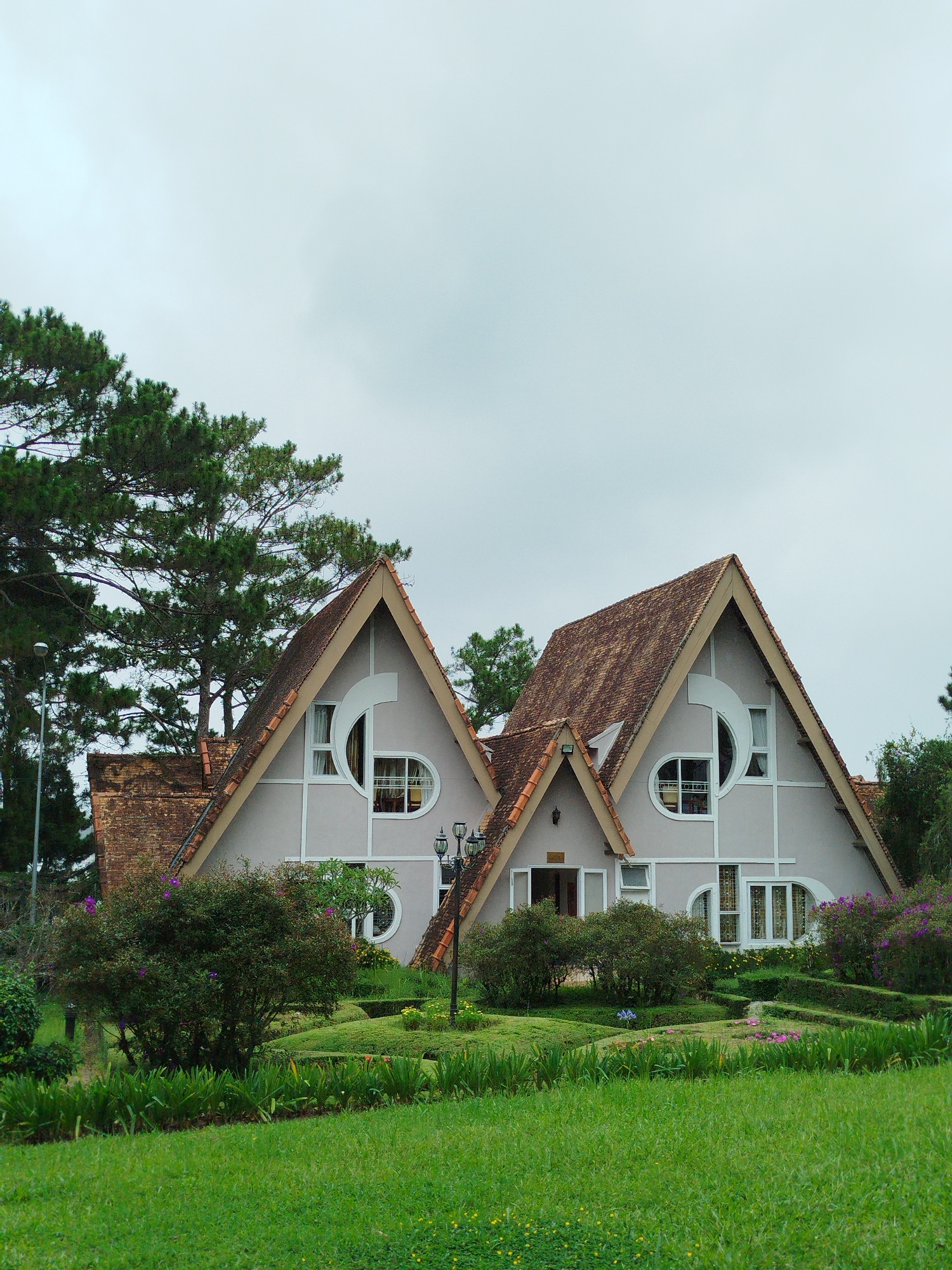 The image shows a pair of adjoining, light-colored, A-frame houses with terracotta tiled roofs. The houses are situated in a well-maintained garden with lush green grass, manicured hedges, and flowering bushes. Tall pine trees are visible behind the houses, and the sky is overcast. The architecture of the houses, particularly the distinctive triangular gables and round windows, is characteristic of many buildings in Da Lat, a city in the Central Highlands of Vietnam known for its French colonial architecture and cooler climate. This style is often referred to as "French Villa" architecture. There are no people visible in the scene. The overall impression is one of a peaceful, somewhat European-style estate set within a verdant landscape.