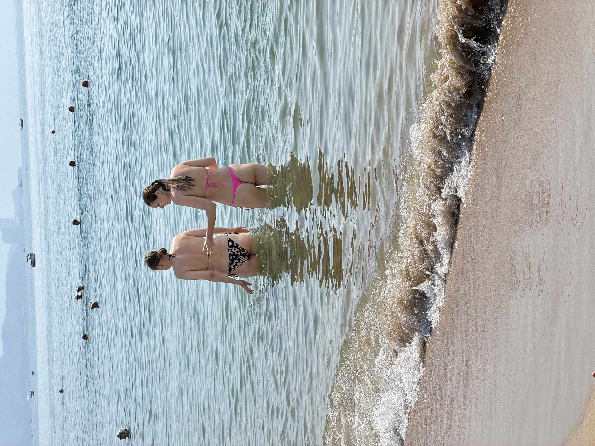 In the city of Nha Trang Ward, Vietnam, two women are standing in the shallow ocean water near the shore. The woman on the left is wearing a black and white patterned bikini, and the woman on the right is wearing a pink bikini. Both women are facing away from the camera. In the background, several other people are visible in the water, and a few boats are on the horizon. The water is calm and clear, with small waves lapping at the sandy shore. The sky is hazy and light blue.