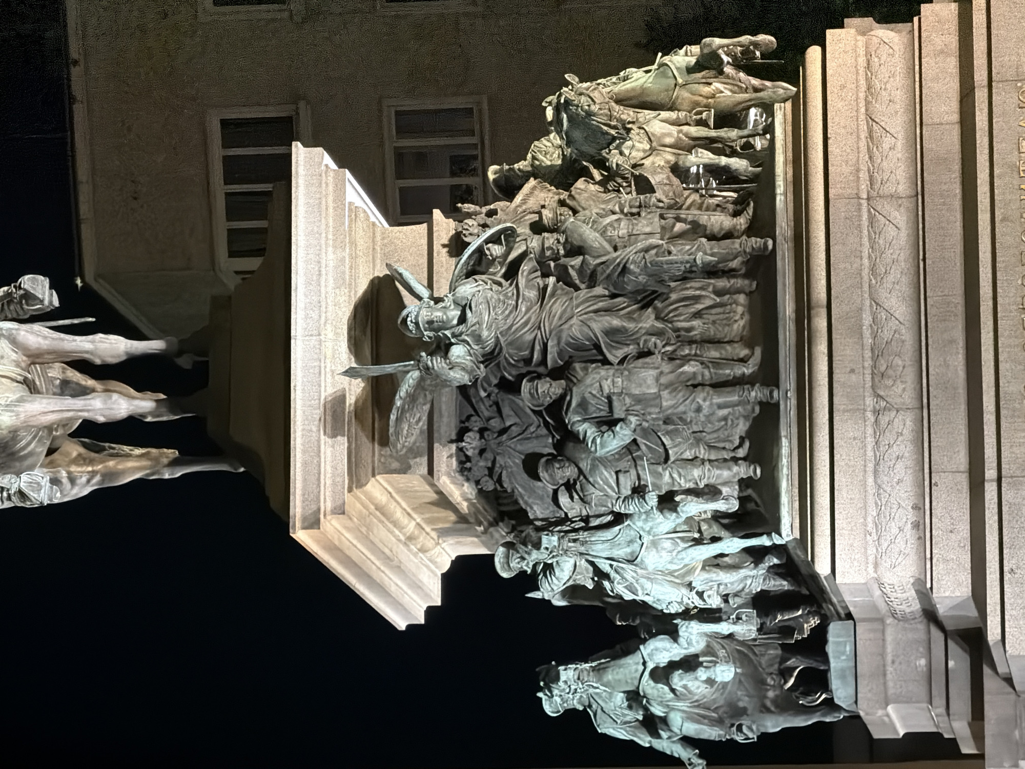 This media file captures a nighttime view of a large, elaborate bronze monument. The monument depicts a dynamic scene of figures, seemingly soldiers and horses, engaged in what appears to be a battle or a march. The composition is dense with many overlapping figures, suggesting a chaotic or intense moment.
The subjects are predominantly bronze sculptures of humans and equines. The human figures are depicted in military attire, some wearing helmets and carrying weapons. The horses are also dramatically posed, with some in mid-gallop. The monument is set against a backdrop of a building, visible in the upper left portion of the image, which has windows and a textured facade. The scene is illuminated by artificial light, casting strong shadows and highlighting the metallic sheen of the sculptures.
The time of day is clearly night due to the darkness in the background and the artificial lighting. There are no indications of weather conditions. The location context provided is Sofia, Bulgaria, suggesting this is a historical monument within the city. There is no visible text in the image that can be clearly deciphered. The overall emotion conveyed by the monument is one of valor, struggle, and perhaps tragedy, as suggested by the active and overlapping poses of the figures. The monument is multifaceted, with different layers of sculpted elements that create a sense of depth and movement.