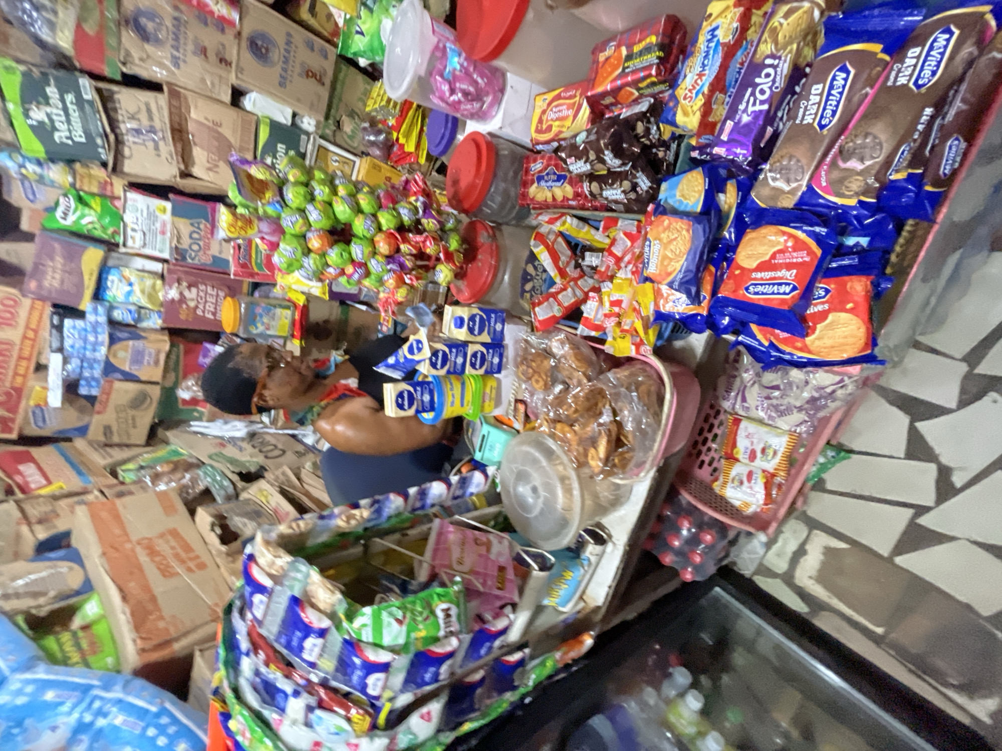 A small shop in Ado, Nigeria, is stocked with a variety of goods. An individual wearing glasses and a cap stands behind a display of packaged snacks and candies, including McVitie's biscuits, lollipops, and other treats. The walls are lined with boxes and packaged goods, creating a crowded environment. A shopping cart filled with various products is positioned in front of the counter.
