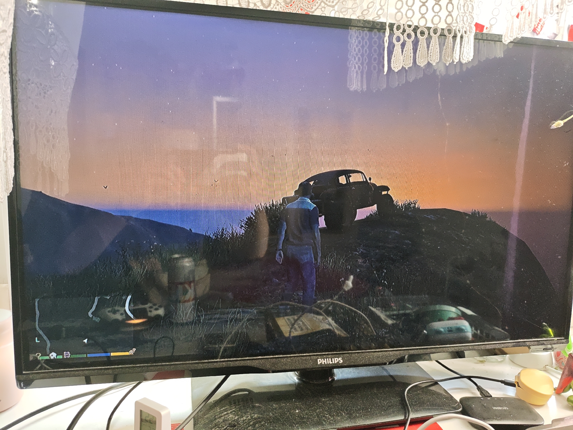 The image shows a computer monitor displaying a video game scene. The game appears to be set in a natural environment with hills and a sunset sky. In the foreground of the game, a person is standing next to a vintage-looking car that is parked on a hilltop. In the background, a body of water is visible. The monitor itself is black and has the "PHILIPS" logo at the bottom center. The screen has reflections of what appear to be lace curtains hanging above it. There are also some smudges and dust on the monitor screen. In front of the monitor, various cables and electronic devices are visible on a desk, including what looks like a small digital clock and a portable charger. The scene is captured indoors, likely in a residential setting. There is no information in the image to suggest any connection to the city of Mława, Poland.